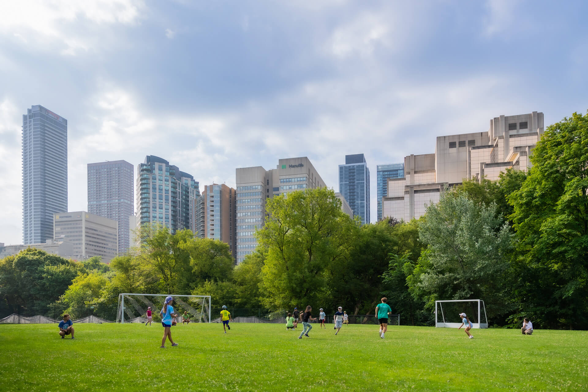 Children play soccer on a grassy field surrounded by trees, with tall city buildings in the background under a partly cloudy sky. Two goalposts are visible on either side of the field.