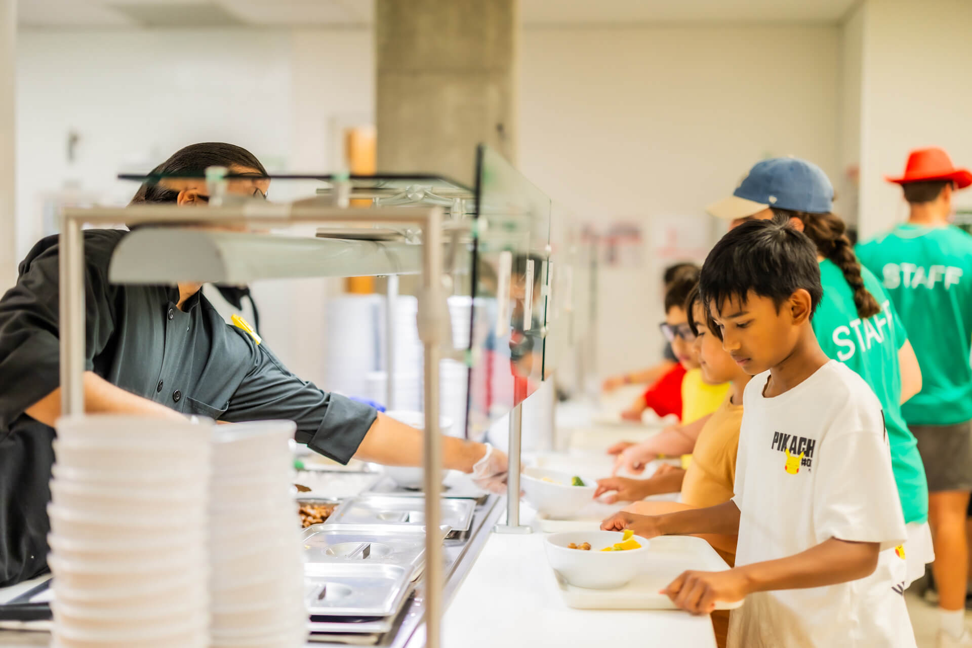 Children stand in line at a cafeteria, holding trays as a staff member serves food behind a glass counter. Other staff, wearing green shirts, assist in the background. The atmosphere appears busy and organized.