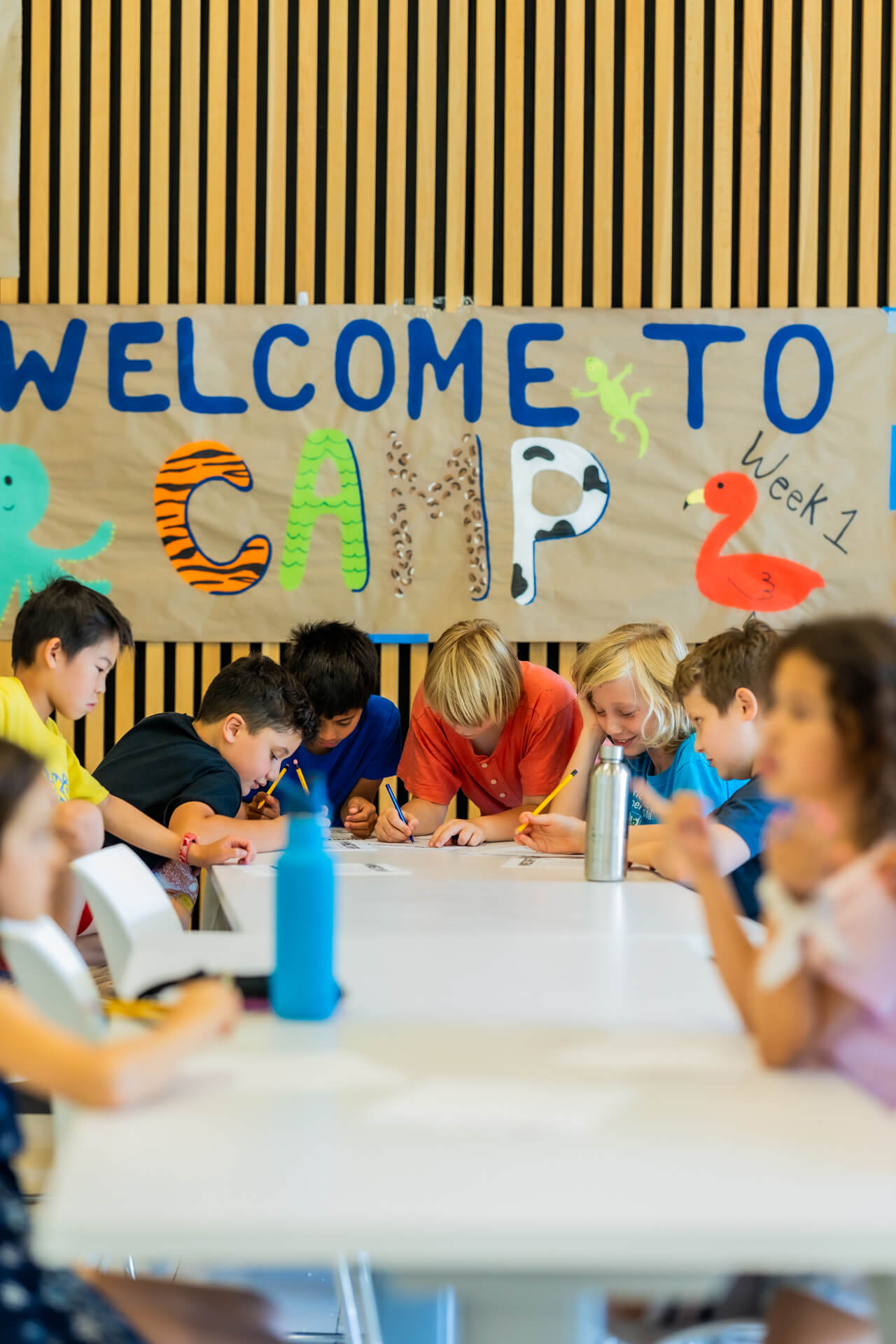A group of children sit at a table drawing or writing, with a colorful "Welcome to Camp" sign decorated with animal shapes hanging behind them.