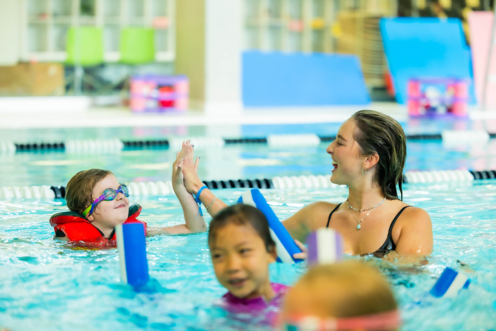 A swim instructor in a pool gives a high-five to a smiling child wearing goggles and a red flotation device, while other children swim nearby during a swimming lesson.