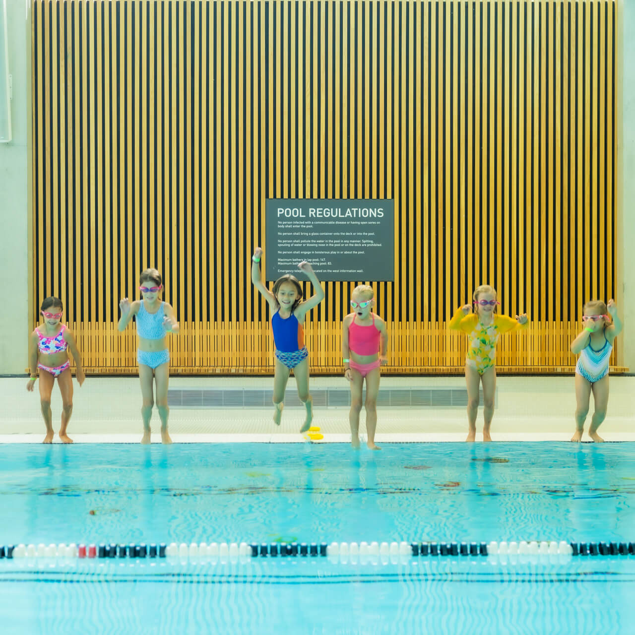 Six young children in colorful swimsuits and goggles are jumping into an indoor swimming pool together, smiling and excited, with a "Pool Regulations" sign visible on the wall behind them.