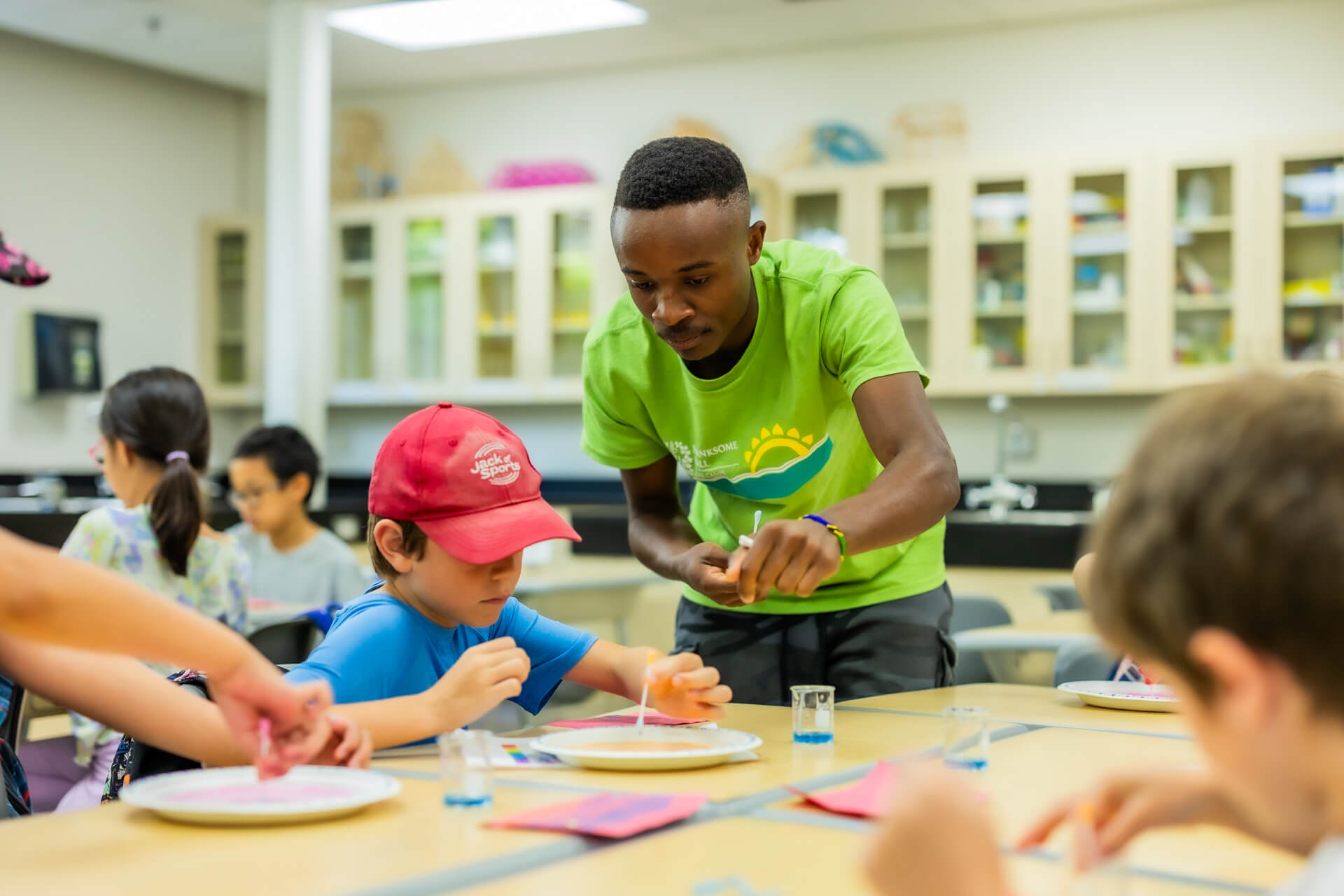 A young adult in a bright green t-shirt helps children with a hands-on science activity at a classroom table. The children focus on the project, using plates and colorful materials. The setting is a well-lit classroom.