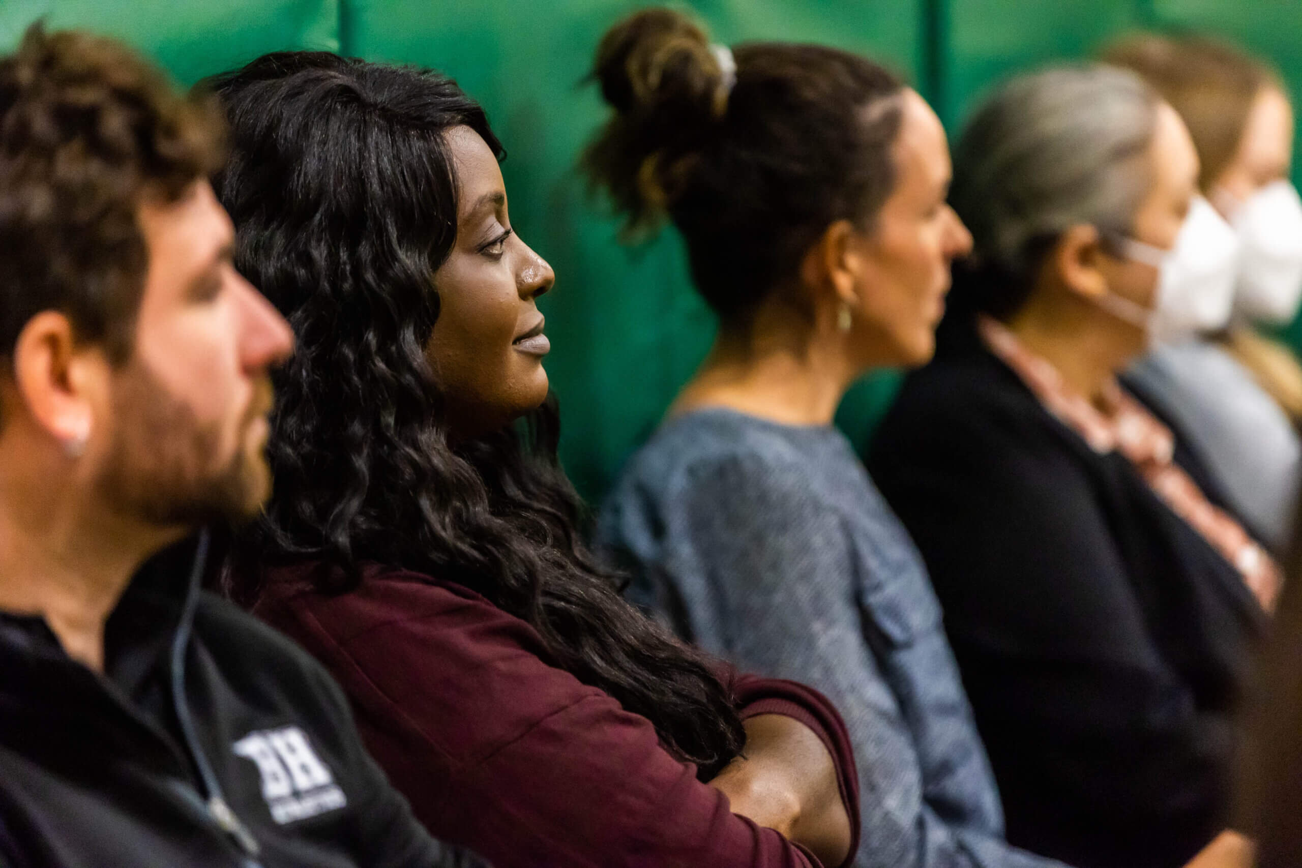 A row of people sitting side by side, attentively watching something off-camera. One person is wearing a mask, while others are focused, with a green padded background behind them.