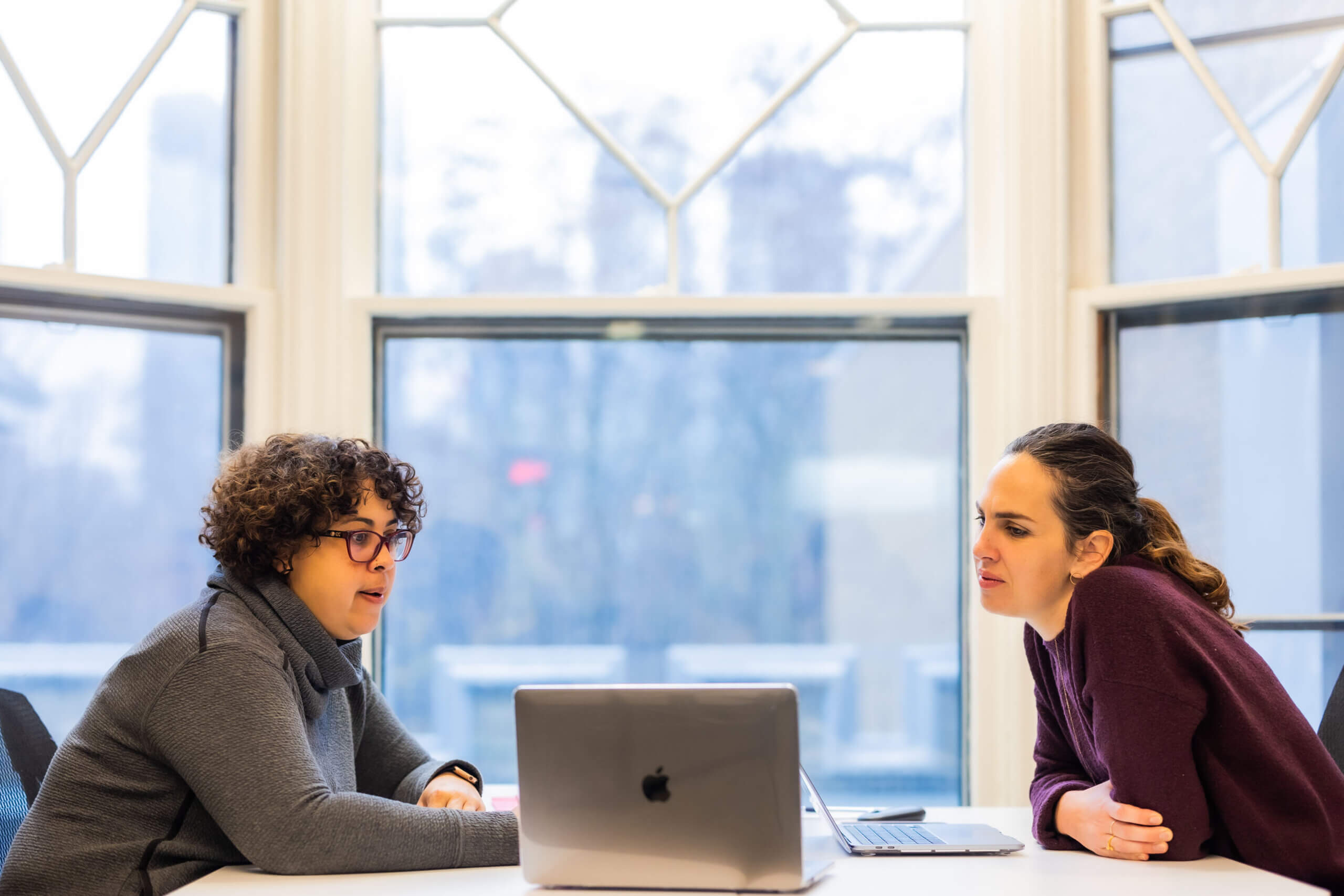 Two people sit across from each other at a table with an open laptop between them, having a discussion. Large windows behind them show a blurred outdoor view.