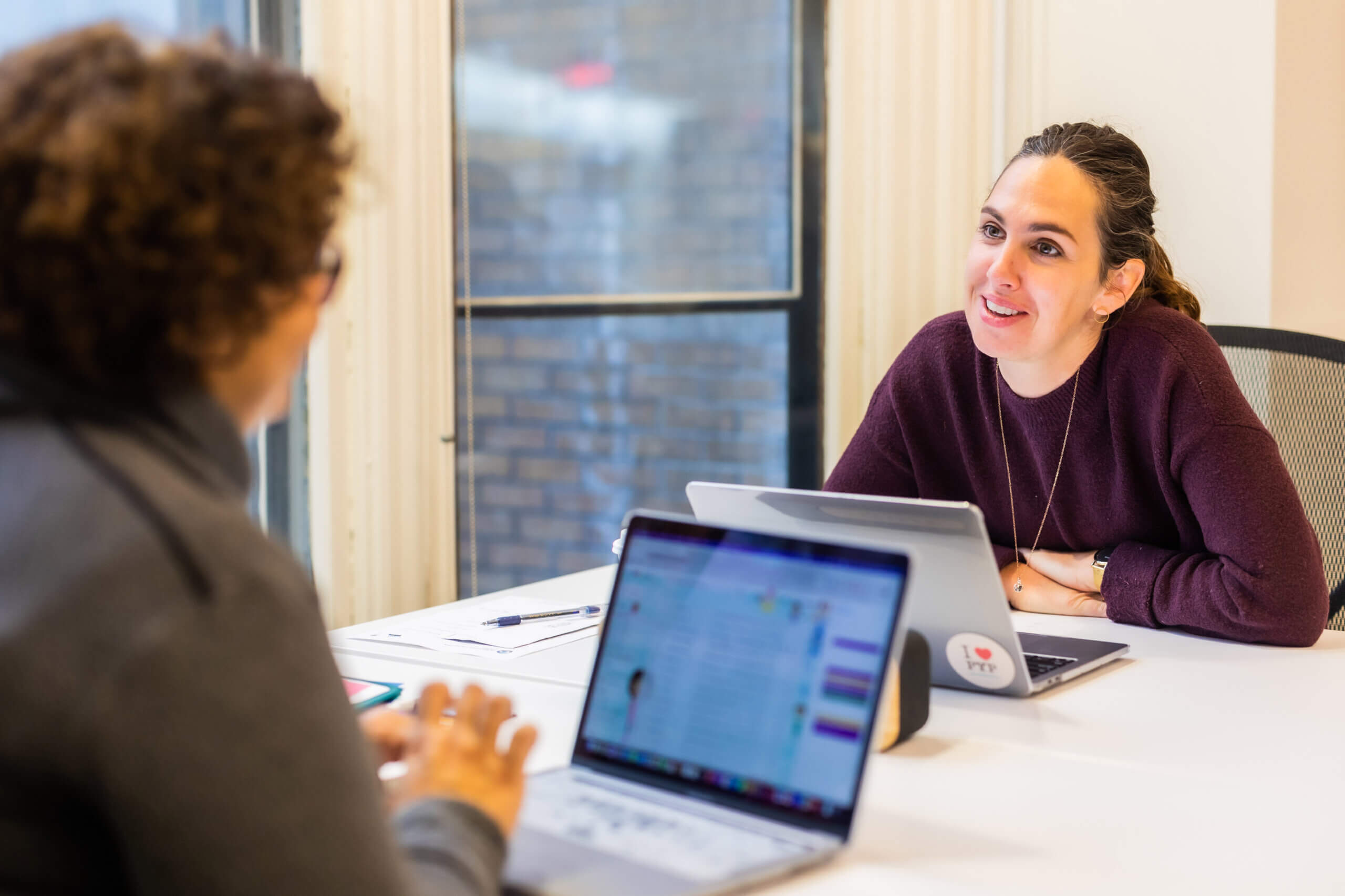 Two women sit across from each other at a table with laptops, having a conversation. One woman is facing the camera and smiling, while the other has her back to the camera. A window is in the background.