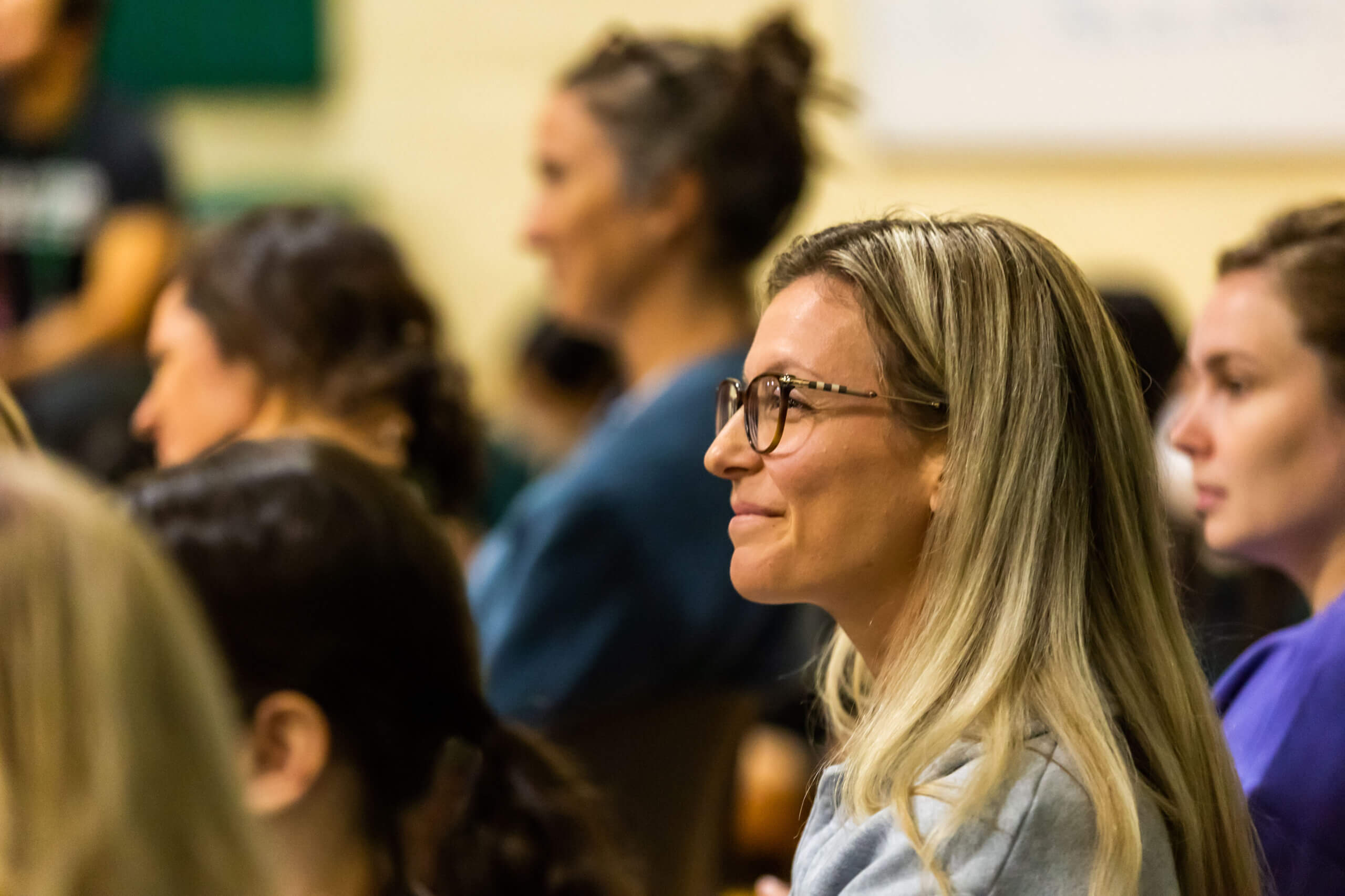 A woman with long blonde hair and glasses is sitting among a group of people, attentively listening and smiling. The background is blurred, focusing on her engaged expression.