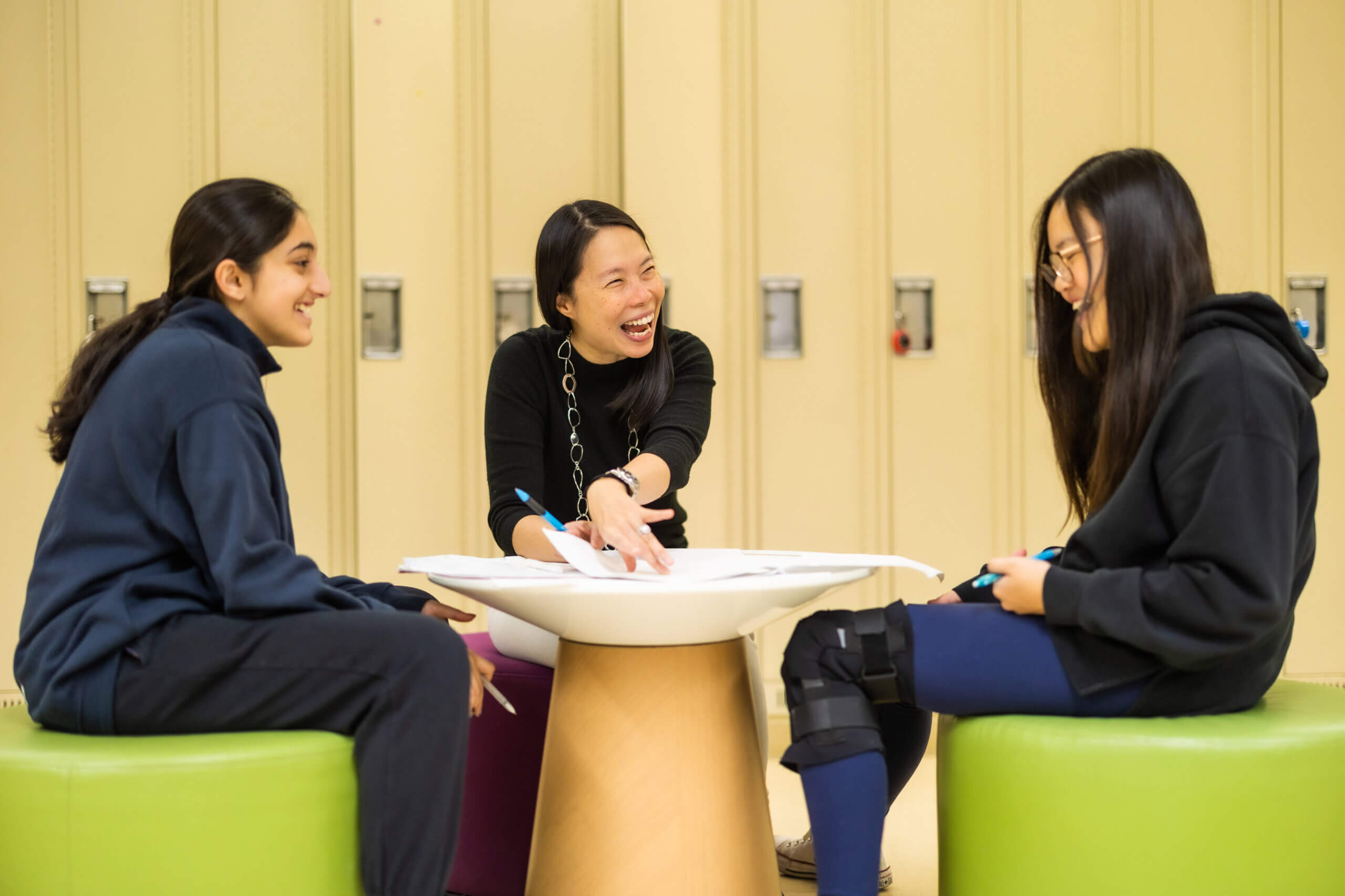 Three young women sit around a small table, smiling and laughing together. They are seated on colorful stools in front of beige lockers, appearing to enjoy a lively conversation or study session.