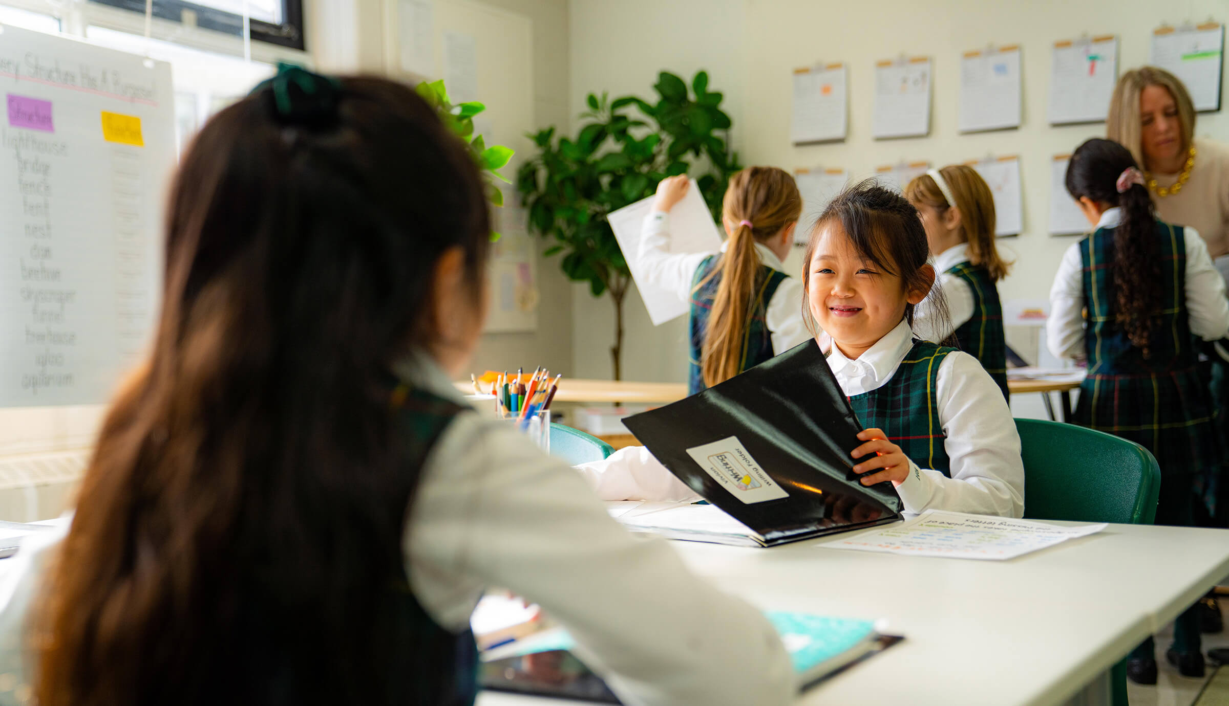 Two young girls in school uniforms sit at a classroom table, smiling and talking. Other students and a teacher are visible in the background. Colorful charts and student work are displayed on the walls.