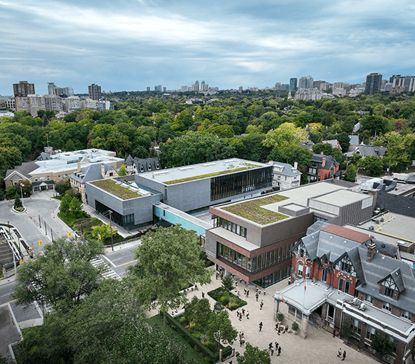 Aerial view of a modern campus with green rooftops, surrounded by trees and city buildings. People walk near the main entrance, and the skyline is visible in the background under a cloudy sky.