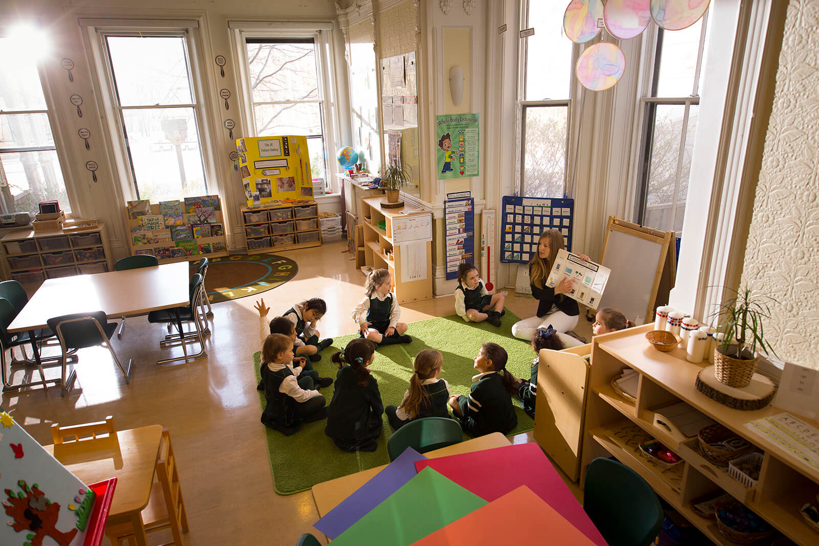 A kindergarten classroom with large windows and natural light; a group of young children and a teacher sit in a circle on a green rug while colorful supplies and learning materials fill the bright, organized room.