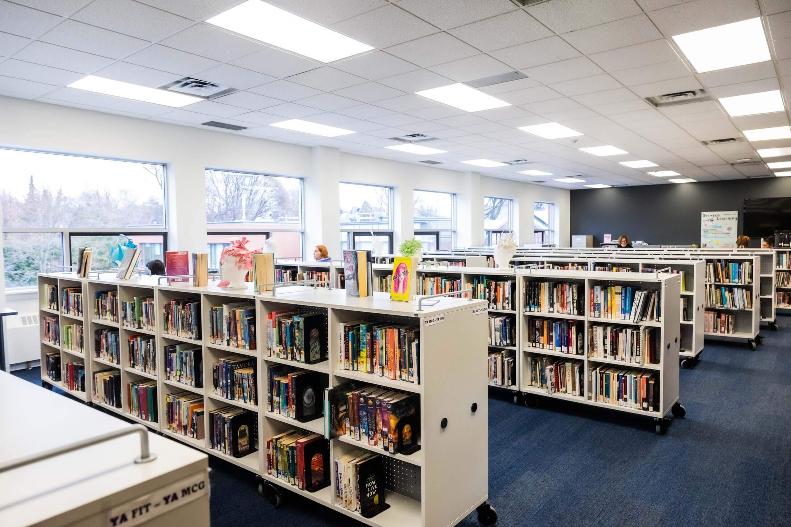 A bright, modern library with large windows, white bookshelves filled with books, and decorations on top. The room has blue carpet, overhead lights, and looks spacious and organized.