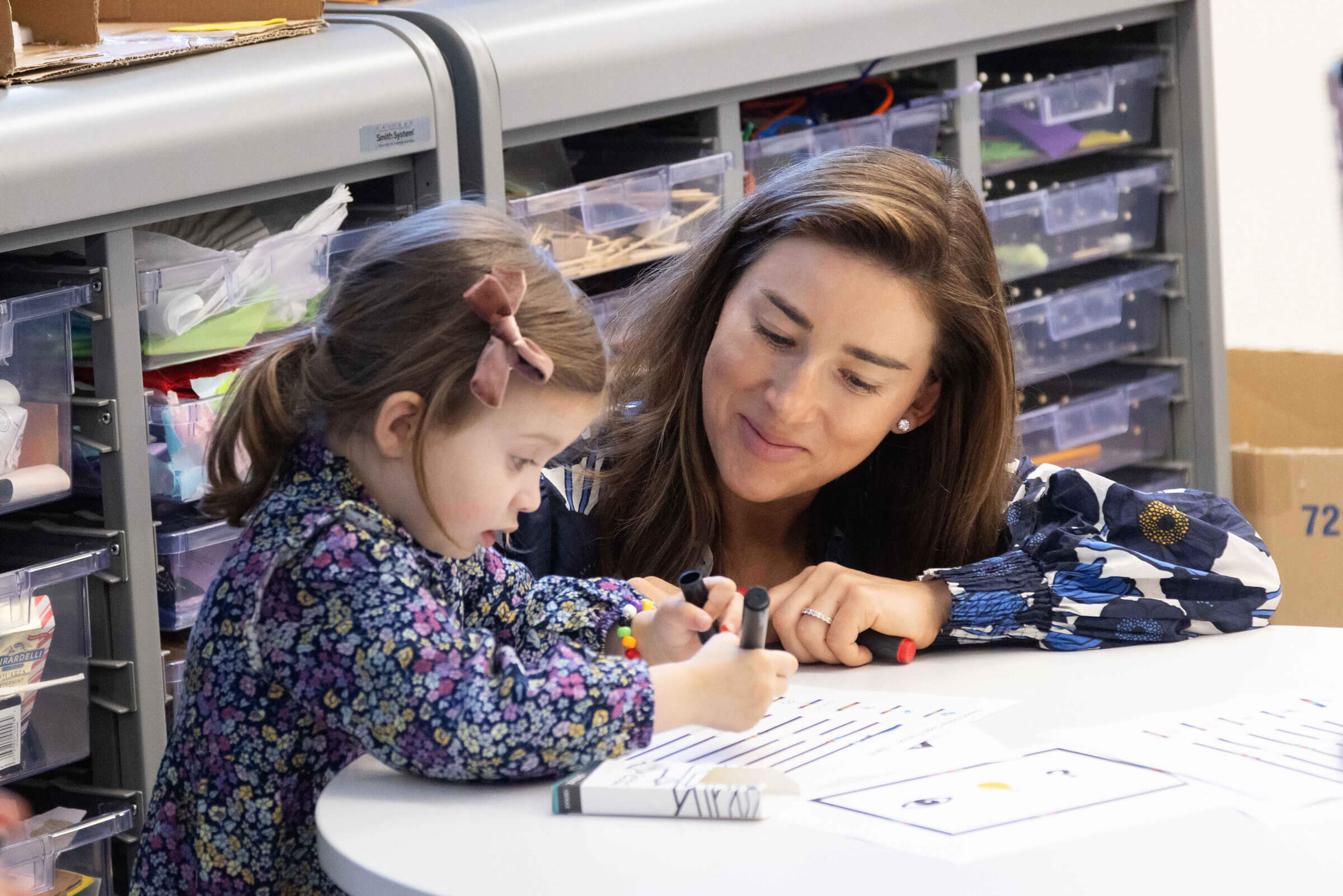 A young girl writes with markers at a table while a woman beside her, possibly a teacher or guardian, watches and smiles supportively. Plastic drawers with art supplies are visible in the background.