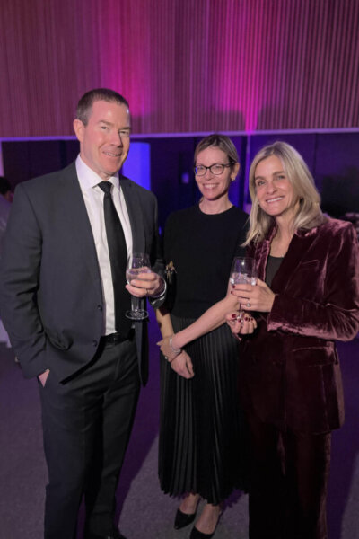 Three adults in formal attire stand together at an indoor event, each holding a drink and smiling at the camera. The background features vertical wooden panels and purple lighting.