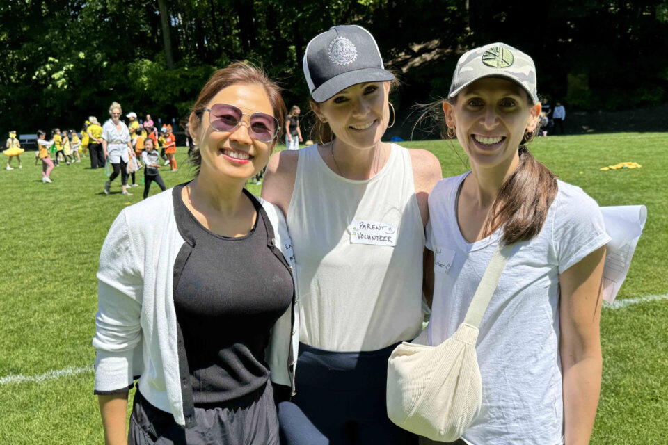 Three women stand together smiling on a sunny grassy field, wearing casual clothes, sunglasses, and hats. Other people and children are visible in the background, suggesting a community or school event outdoors.