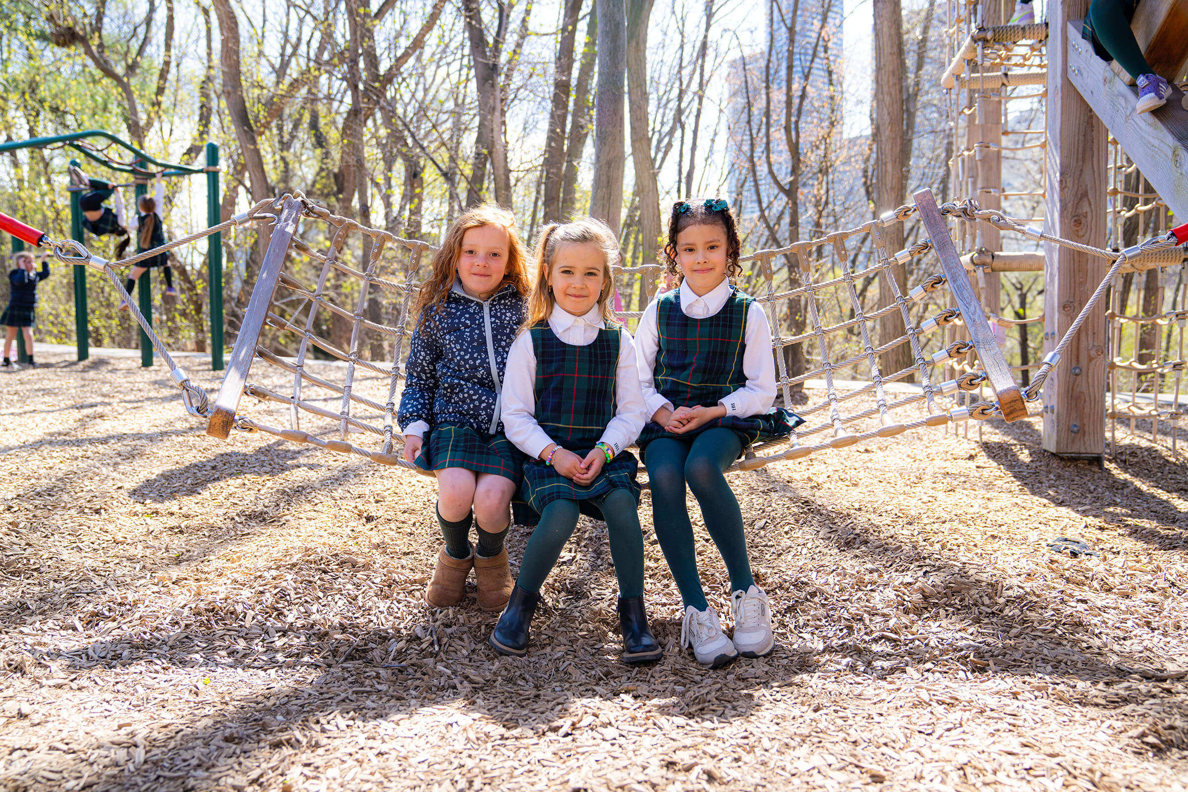 Three young girls in school uniforms sit together on a rope swing in a sunny playground surrounded by trees, with other children playing in the background.
