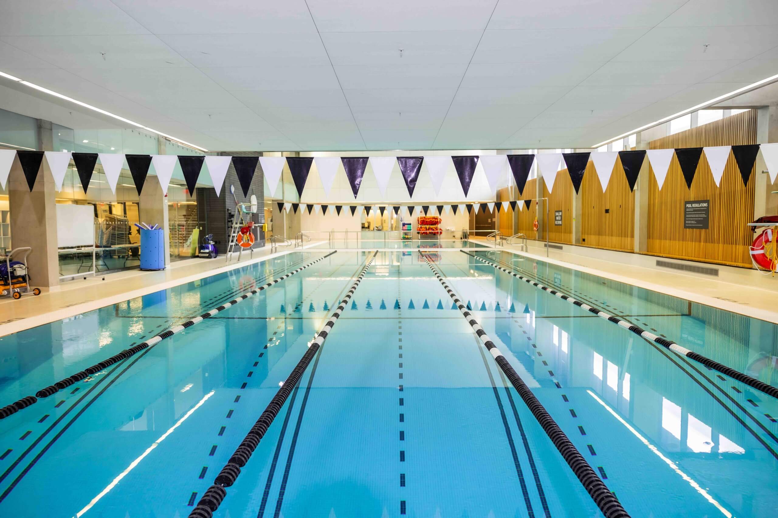 Indoor swimming pool with clear blue water, marked lane lines, and black-and-white triangular flags hanging above. The pool is surrounded by bright lights and modern wood and glass walls. No people are present.