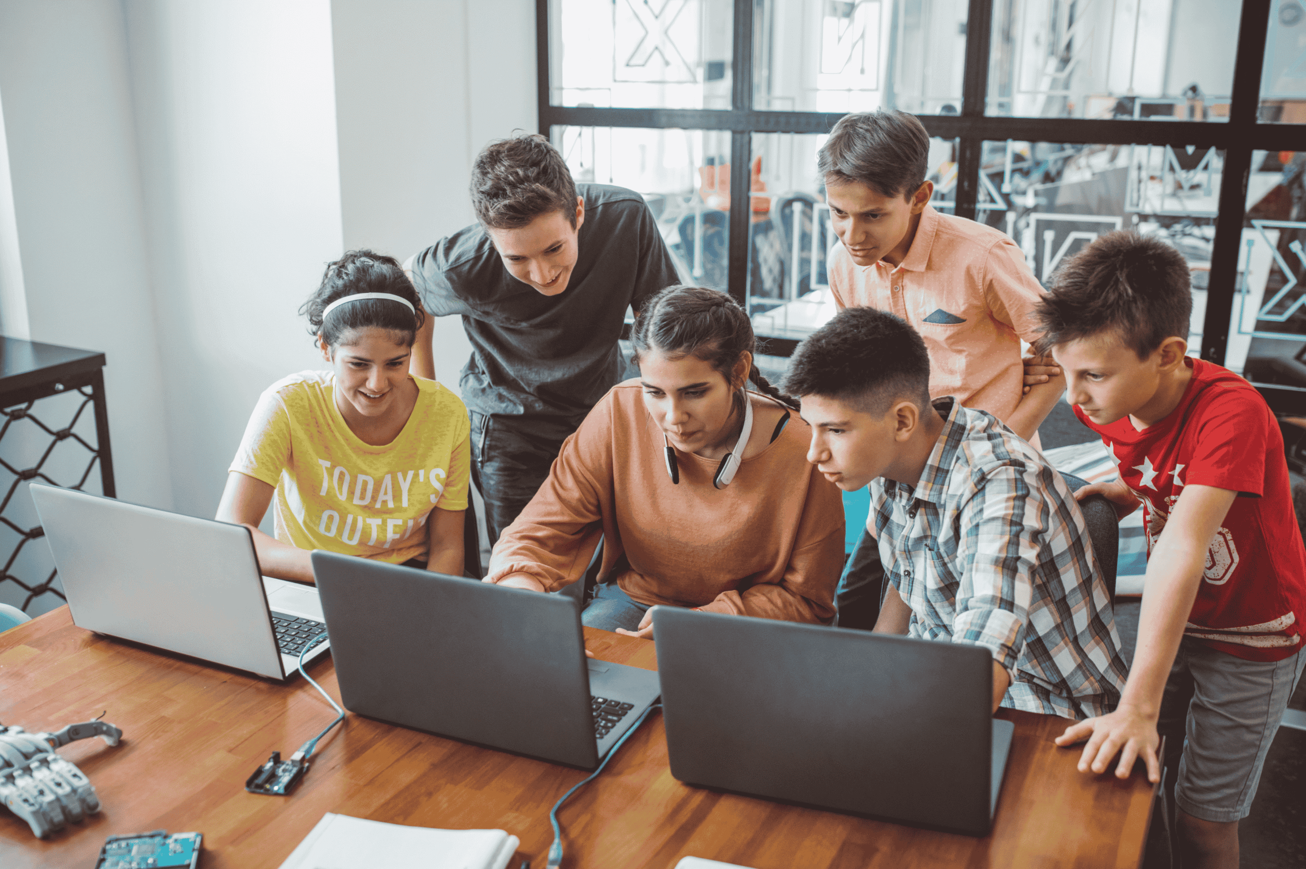 A group of six teenagers, three boys and three girls, gather around a table with laptops, collaborating and looking intently at one screen in a bright, modern classroom.