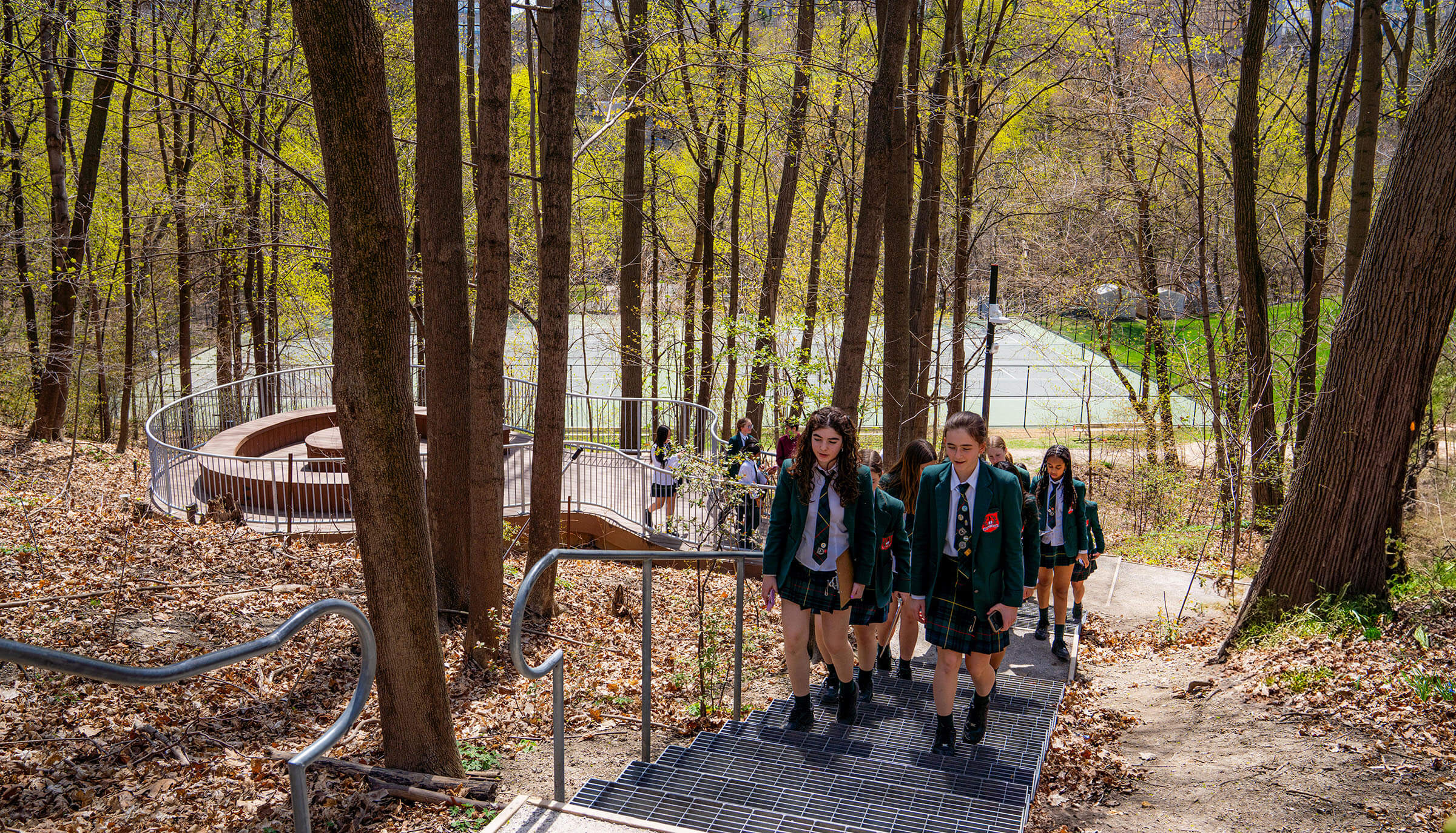 A group of students in school uniforms walk up outdoor metal stairs through a wooded area in early spring, with tennis courts visible in the background.