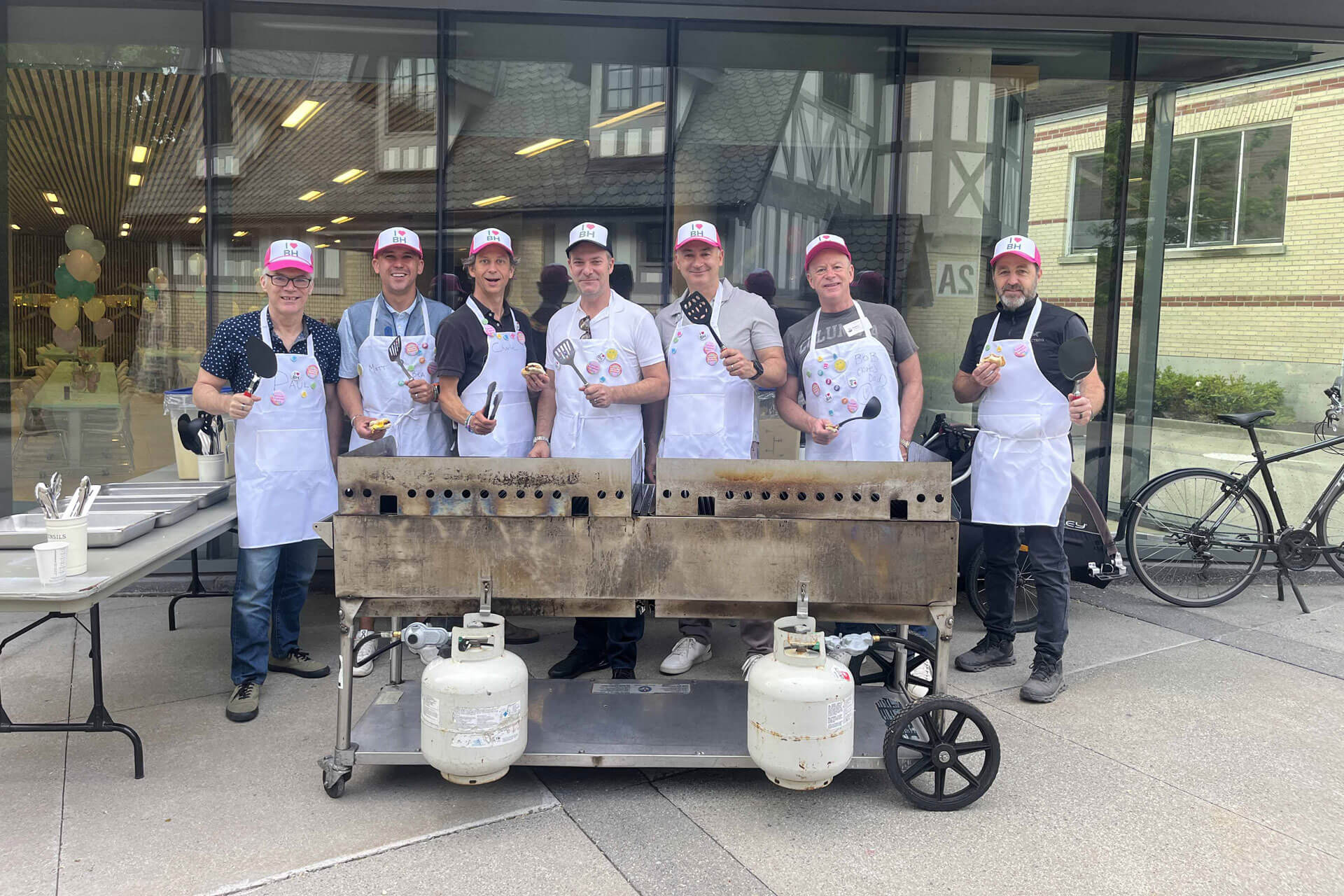Seven people wearing aprons and hats stand behind a large outdoor grill, smiling and holding grilling tools and food. They are in front of a building with large windows and a bicycle nearby.