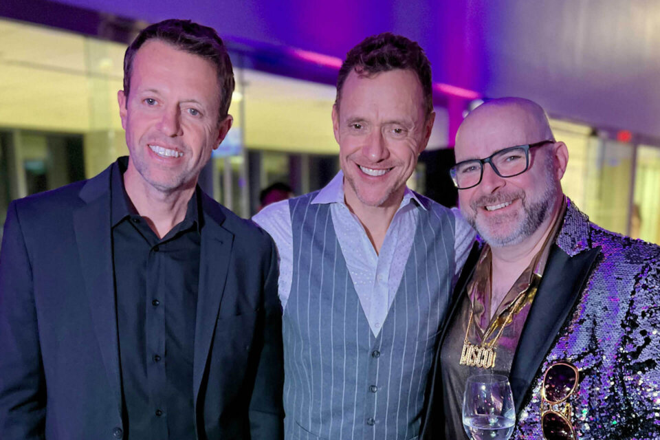 Three men pose together at a formal event. One wears a black suit, another a gray pinstripe vest over a white shirt, and the third wears a shiny sequin jacket, glasses, gold chains, and holds a wine glass. The lighting is purple and blue.
