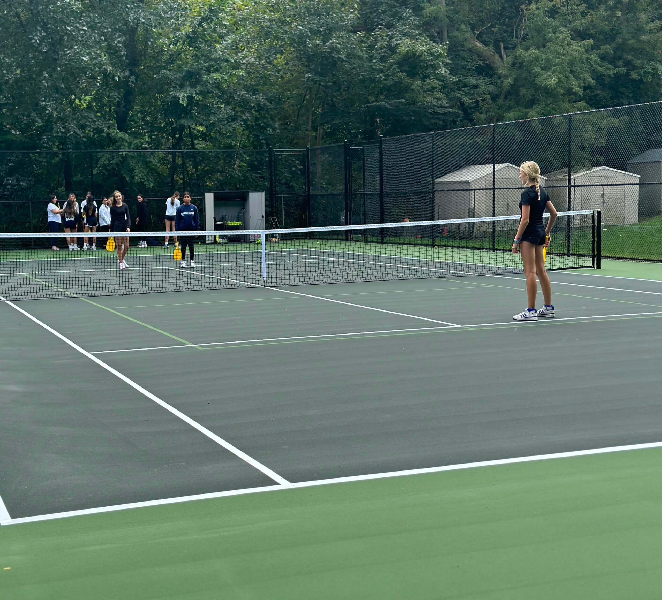 A woman in sportswear stands ready on a tennis court while three people watch from outside the fence in the background, surrounded by trees and sheds.