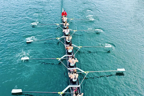 A rowing team of eight people rows in sync on calm blue water, guided by a coxswain at the front of the long narrow boat. The image is taken from above.