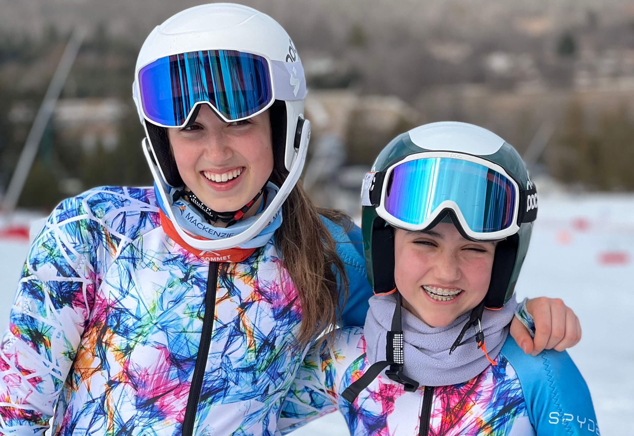 Two young skiers in colorful ski jackets and helmets smile at the camera, standing arm in arm on a snowy slope with blurred trees and buildings in the background.