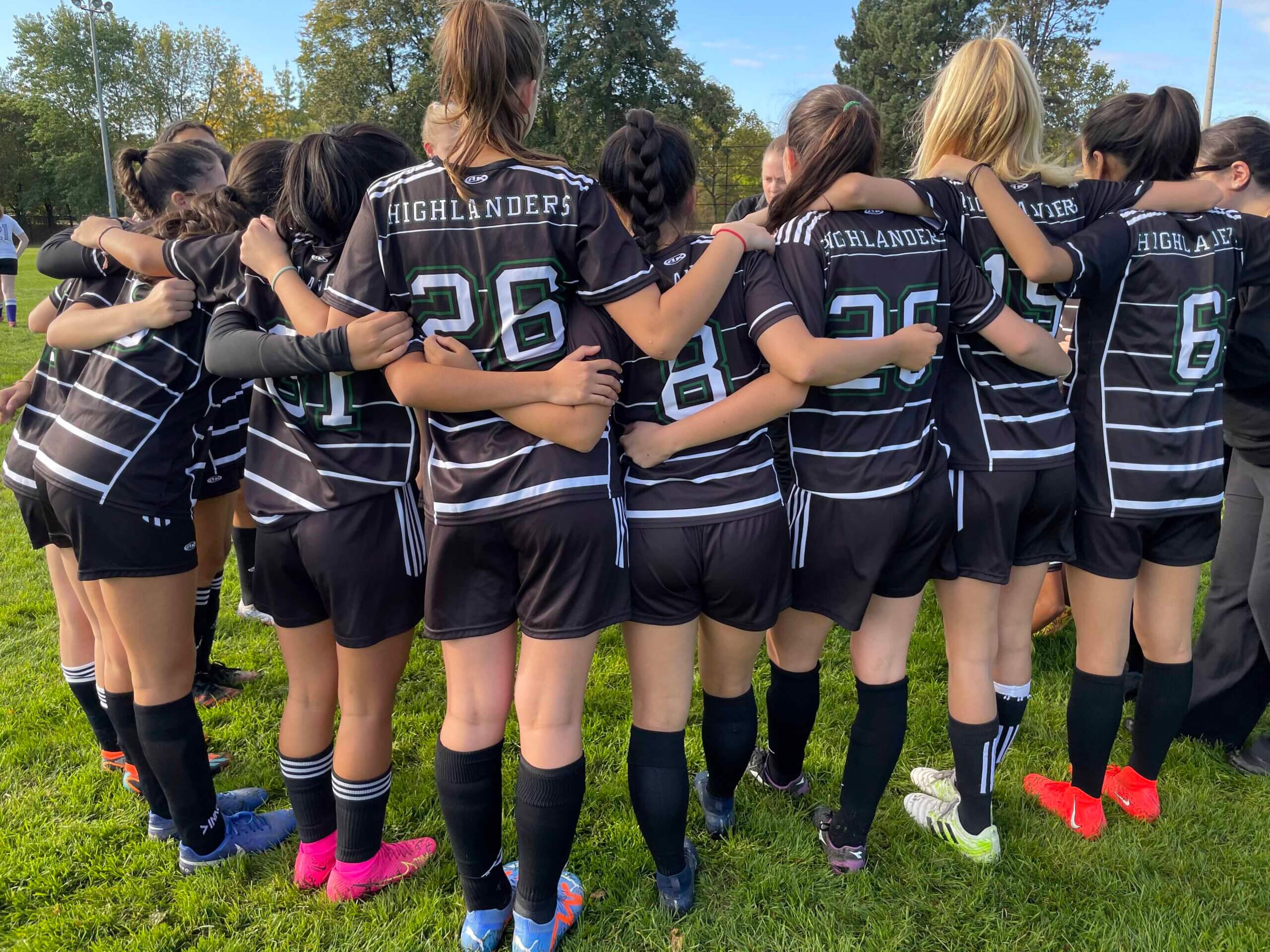 A girls' soccer team in black uniforms with "Highlanders" on their jerseys stands in a huddle on a grassy field, arms around each other, preparing for a game. Trees and blue sky are visible in the background.