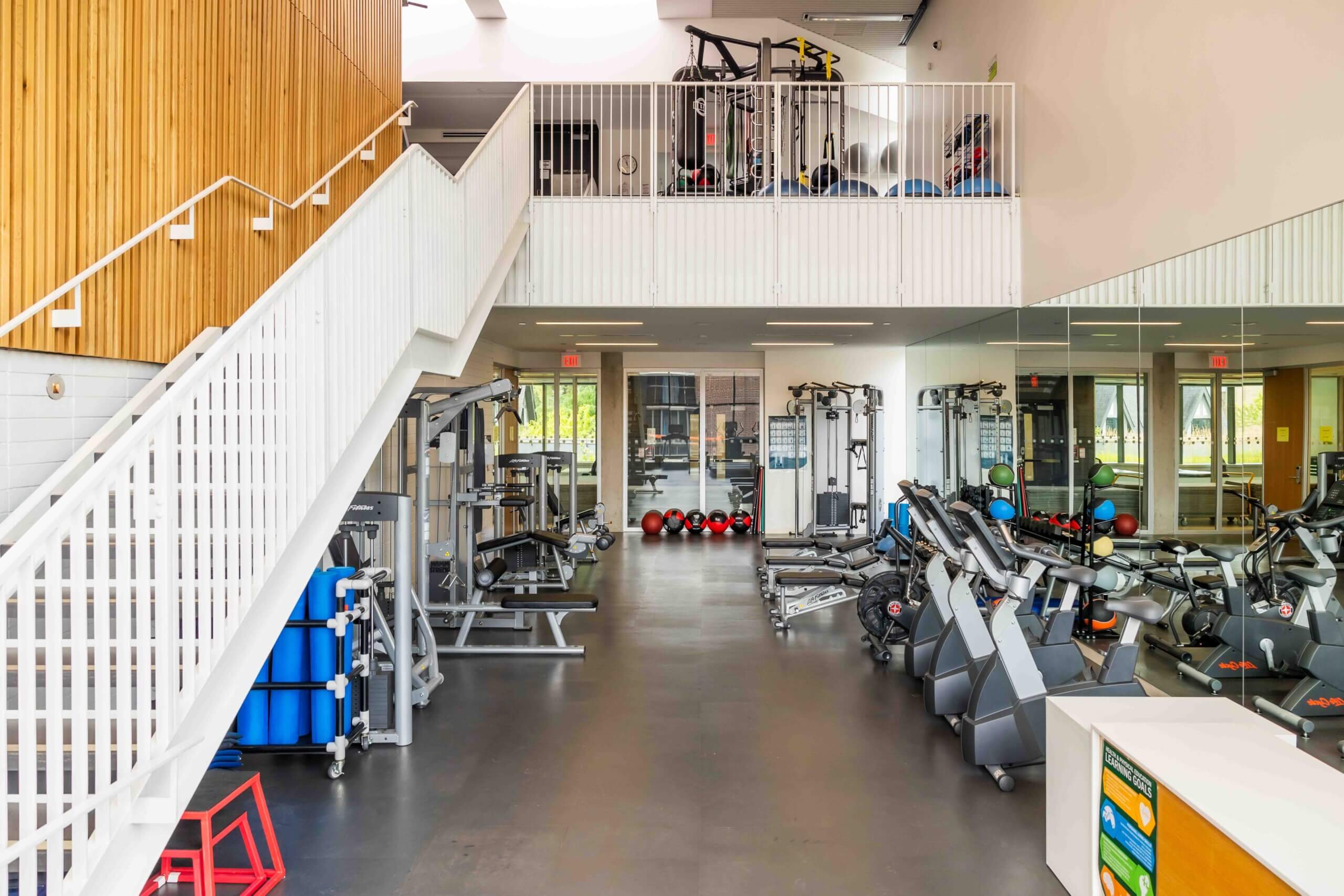 A modern gym with various exercise machines, free weights, and cardio equipment. There is a white staircase on the left, large mirrors on the right, and natural light coming in from windows and skylights.