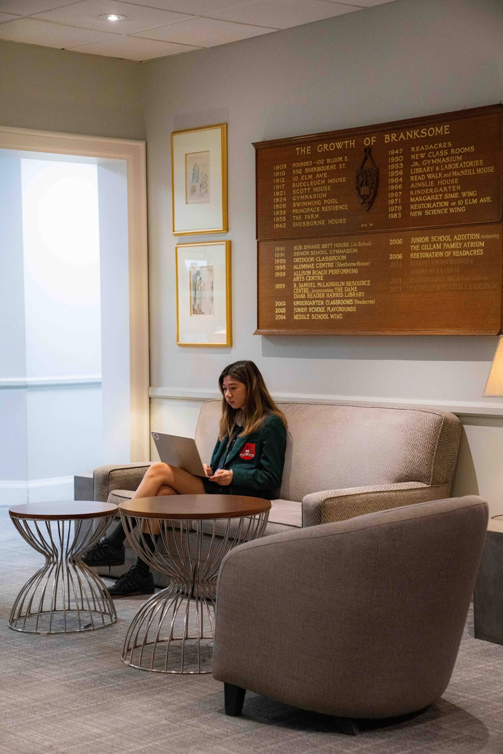 A young woman sits on a sofa using a laptop in a lounge area with modern furniture. Behind her is a wooden plaque listing milestones for "The Growth of Branksome" and framed artwork on the wall.