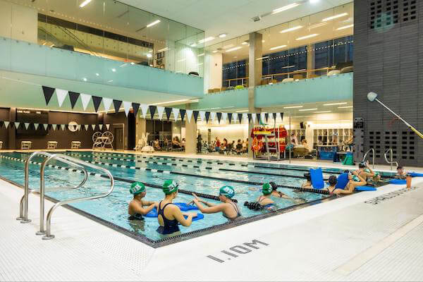 Indoor swimming pool with swimmers practicing along the edge, wearing green swim caps and using kickboards. In the background, people are seated and walking in a brightly lit, modern facility.