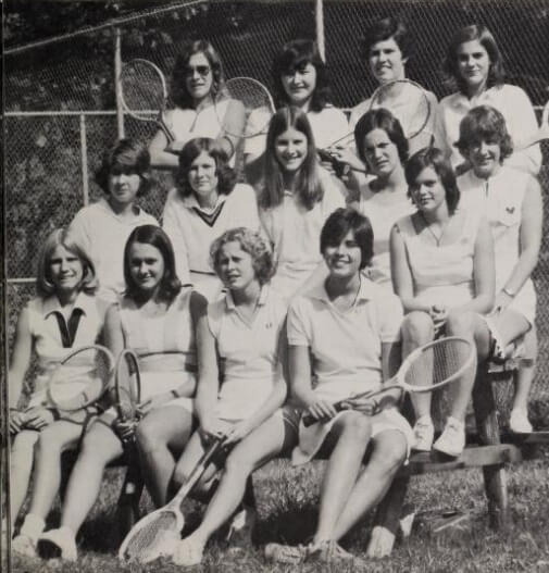 A black-and-white photo of a girls’ tennis team, with 14 young women posing outdoors in front of a chain-link fence. Some are seated holding tennis rackets, while others stand behind them, all wearing tennis uniforms.