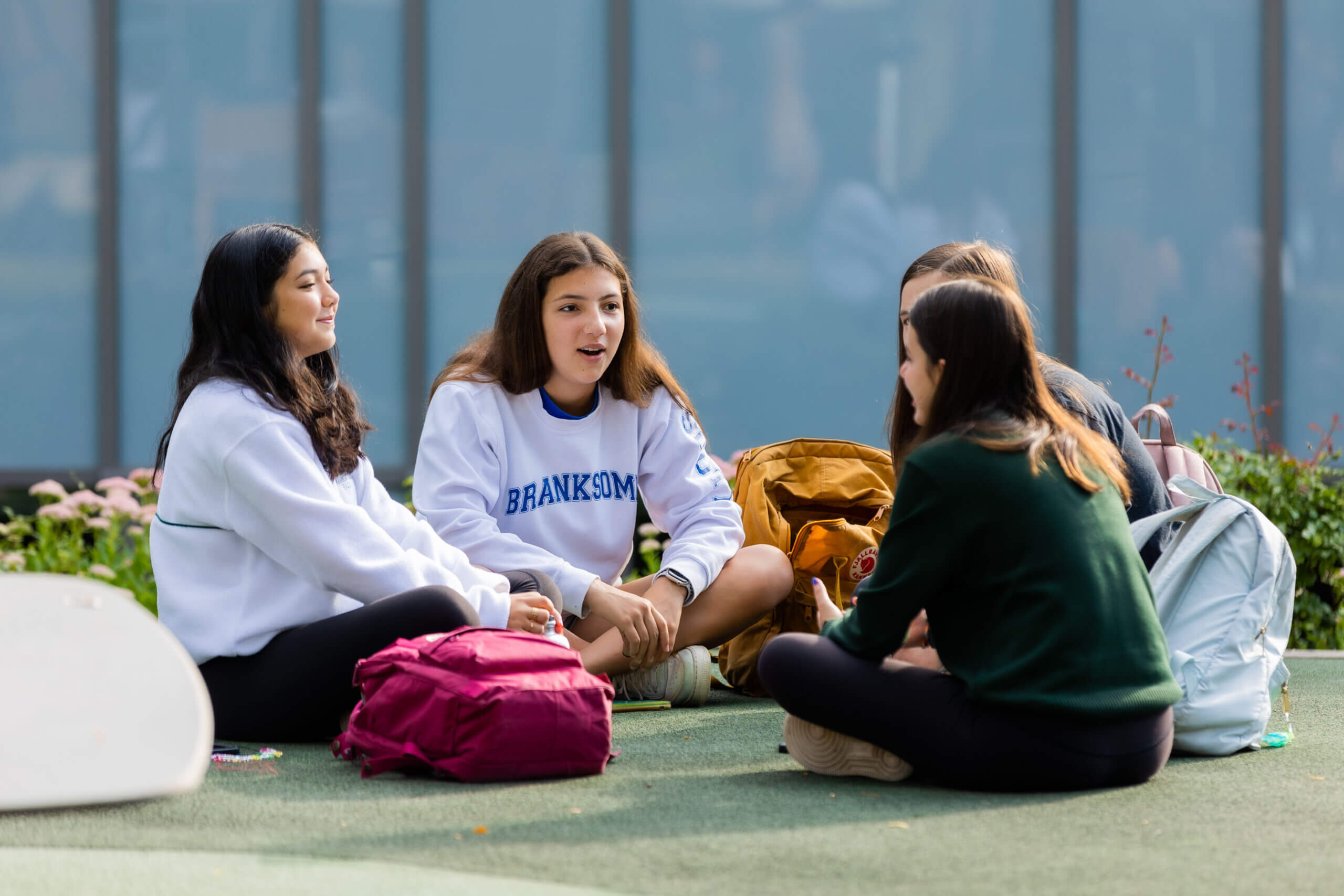 Four teenage girls sit in a circle on the grass outside, talking and smiling, with backpacks around them. One wears a white "Branksome" sweatshirt. A glass building is visible in the background.
