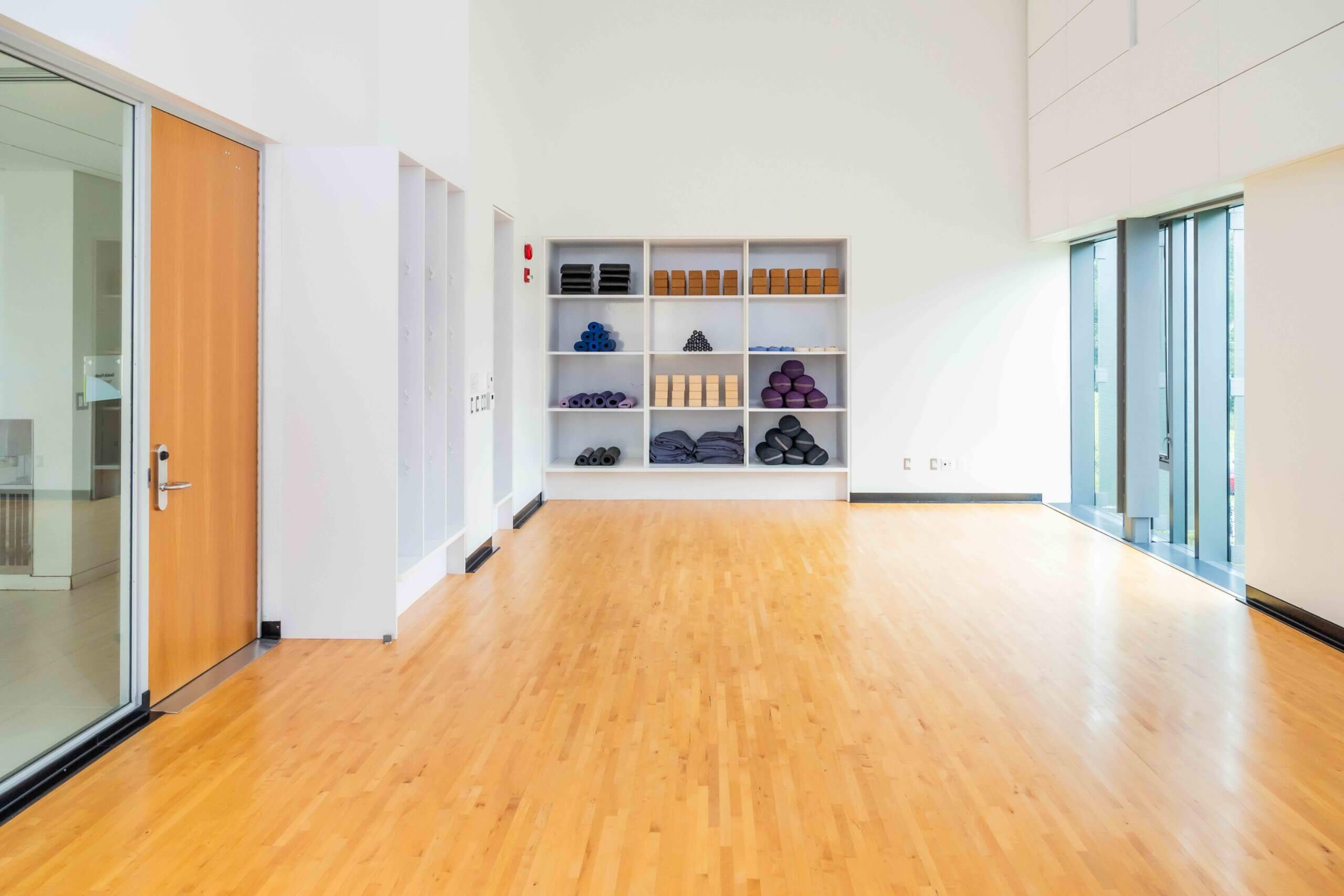 Bright, spacious studio with a wood floor, white walls, glass doors, and large windows. Shelves at the back hold neatly stacked yoga mats, blocks, bolsters, and blankets.