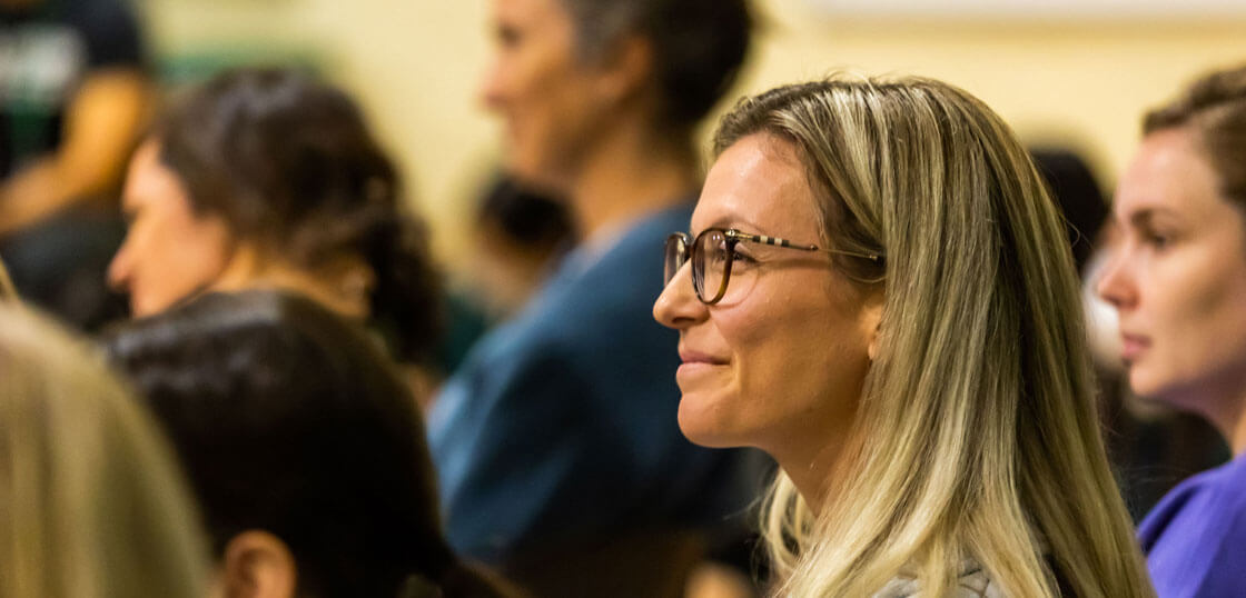 A woman with long blonde hair and glasses smiles while sitting among a group of people, all attentively watching something out of frame.