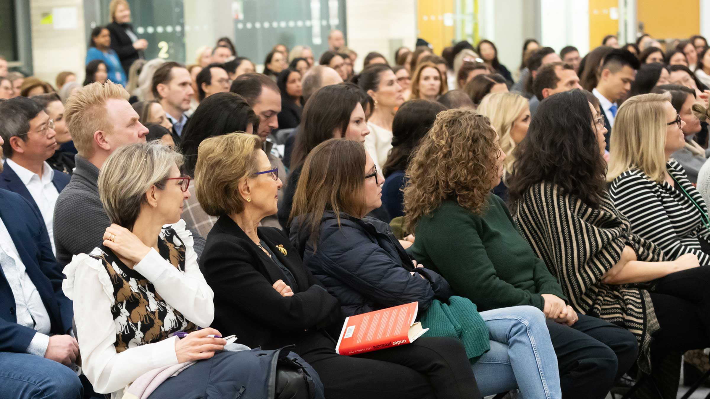 A large audience of men and women sits closely together indoors, attentively watching an event. Most people are facing forward; some hold notebooks and wear business or casual attire.
