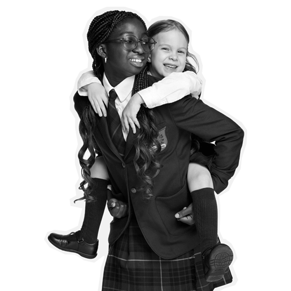 Two girls in school uniforms smile as one gives the other a piggyback ride. Both wear blazers, ties, and skirts. The younger girl, with light hair, hugs the older girl, who has long braids and glasses. The background is plain white.
