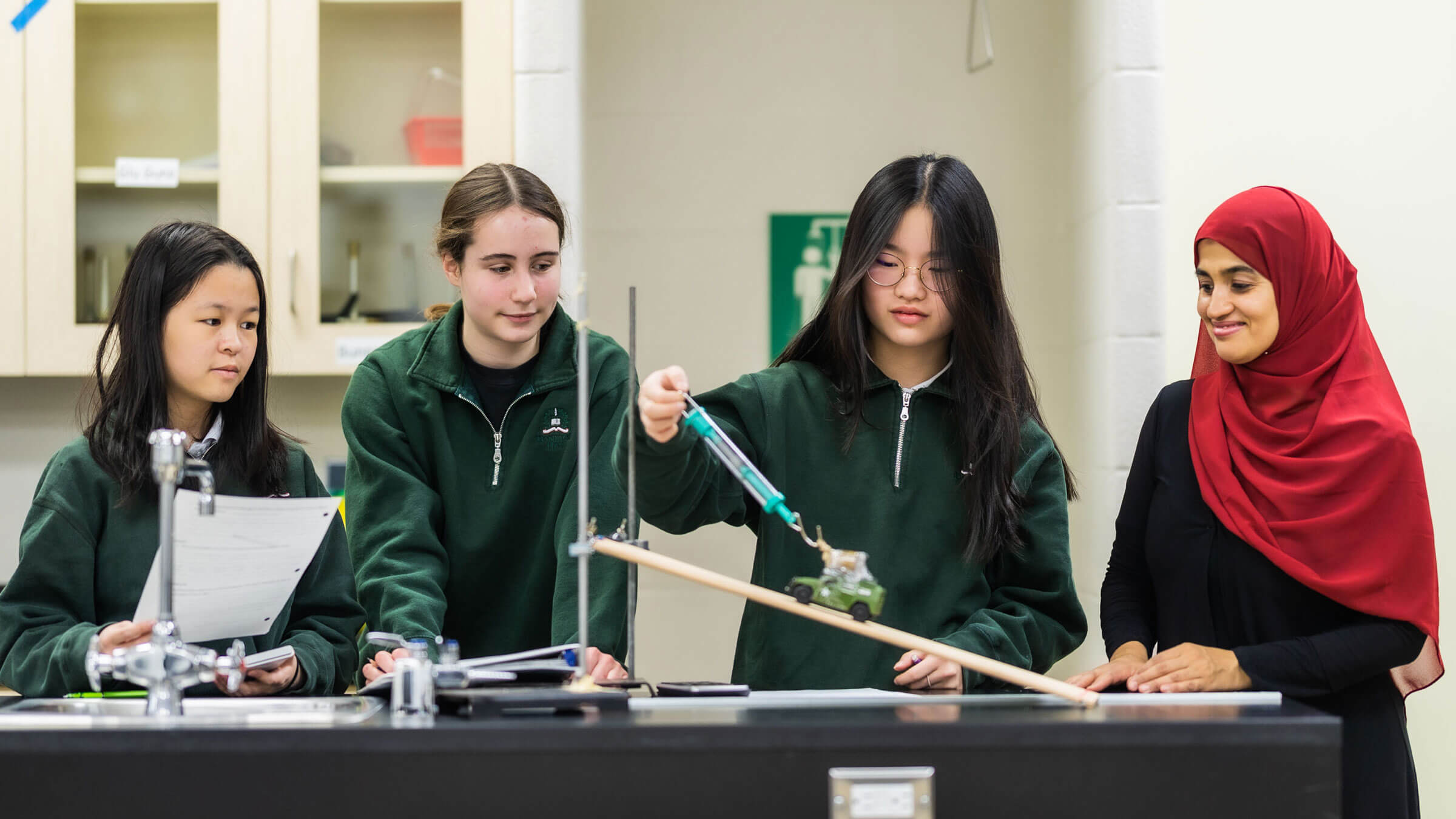 Four young women in a science lab work together on an experiment with a ramp and a small cart, supervised by a teacher wearing a red hijab. They appear focused and engaged in the activity.