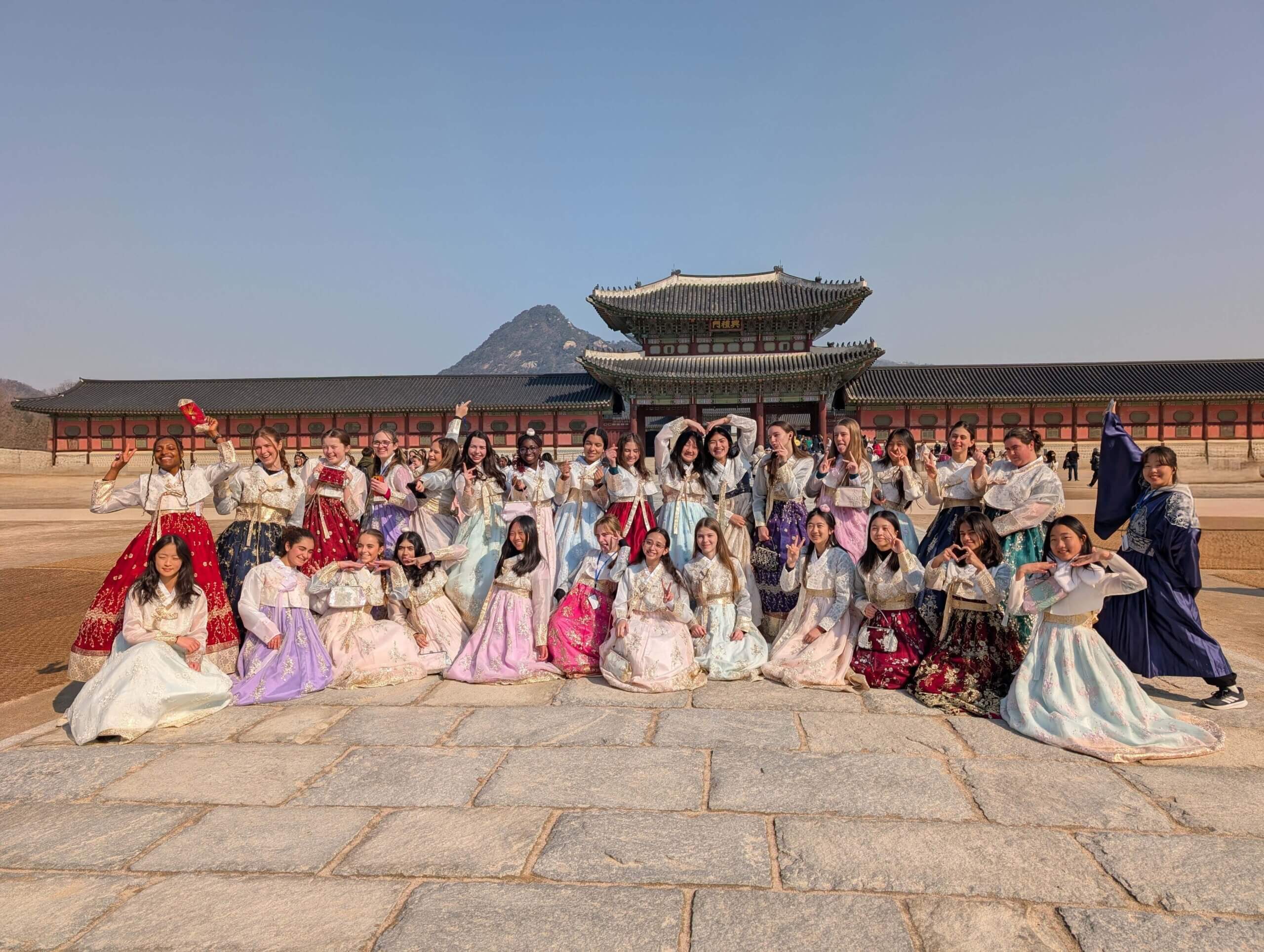 A large group of people pose together in colorful traditional Korean hanbok outfits in front of a historic palace building, with mountains visible in the background under a clear sky.
