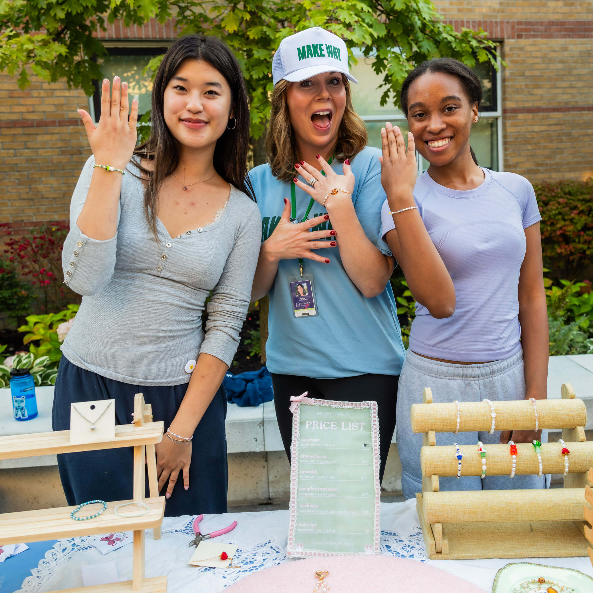 Three young women smile and show off beaded bracelets at an outdoor jewelry booth. A price list and displays of handmade jewelry are on the table in front of them, with green trees and a building in the background.