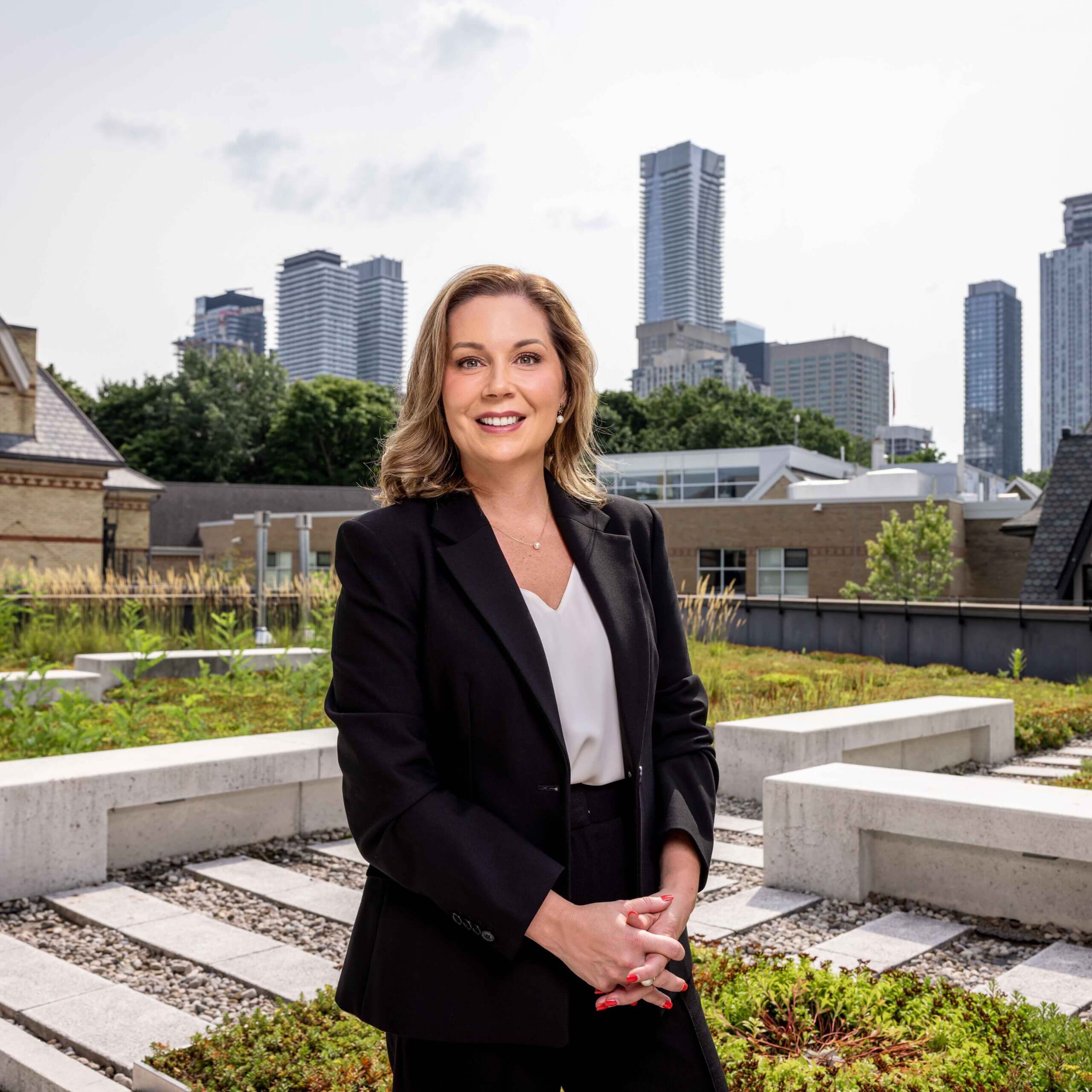 A woman in a black suit stands on a rooftop garden, smiling at the camera. Modern city skyscrapers and buildings are visible in the background under a partly cloudy sky.