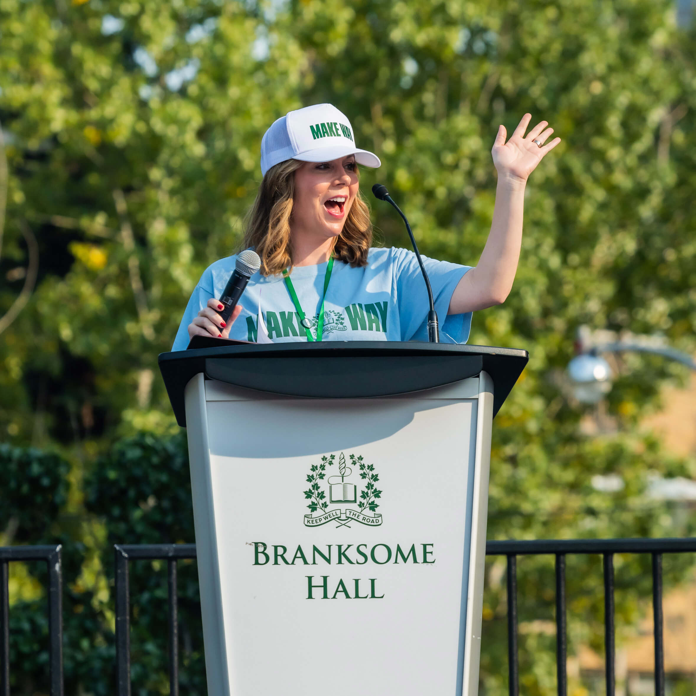 A woman wearing a "MAKE WAY" hat and T-shirt speaks enthusiastically at an outdoor podium labeled "Branksome Hall," holding a microphone with one hand raised and trees visible in the background.
