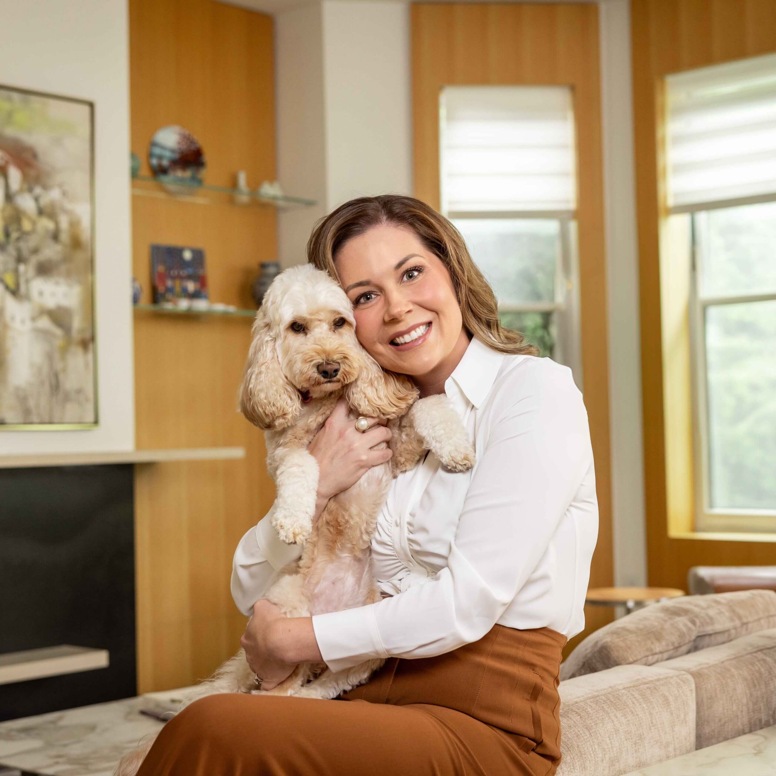 A woman in a white blouse and brown skirt sits on a sofa, smiling and hugging a small, fluffy, light-colored dog in a bright, modern living room.