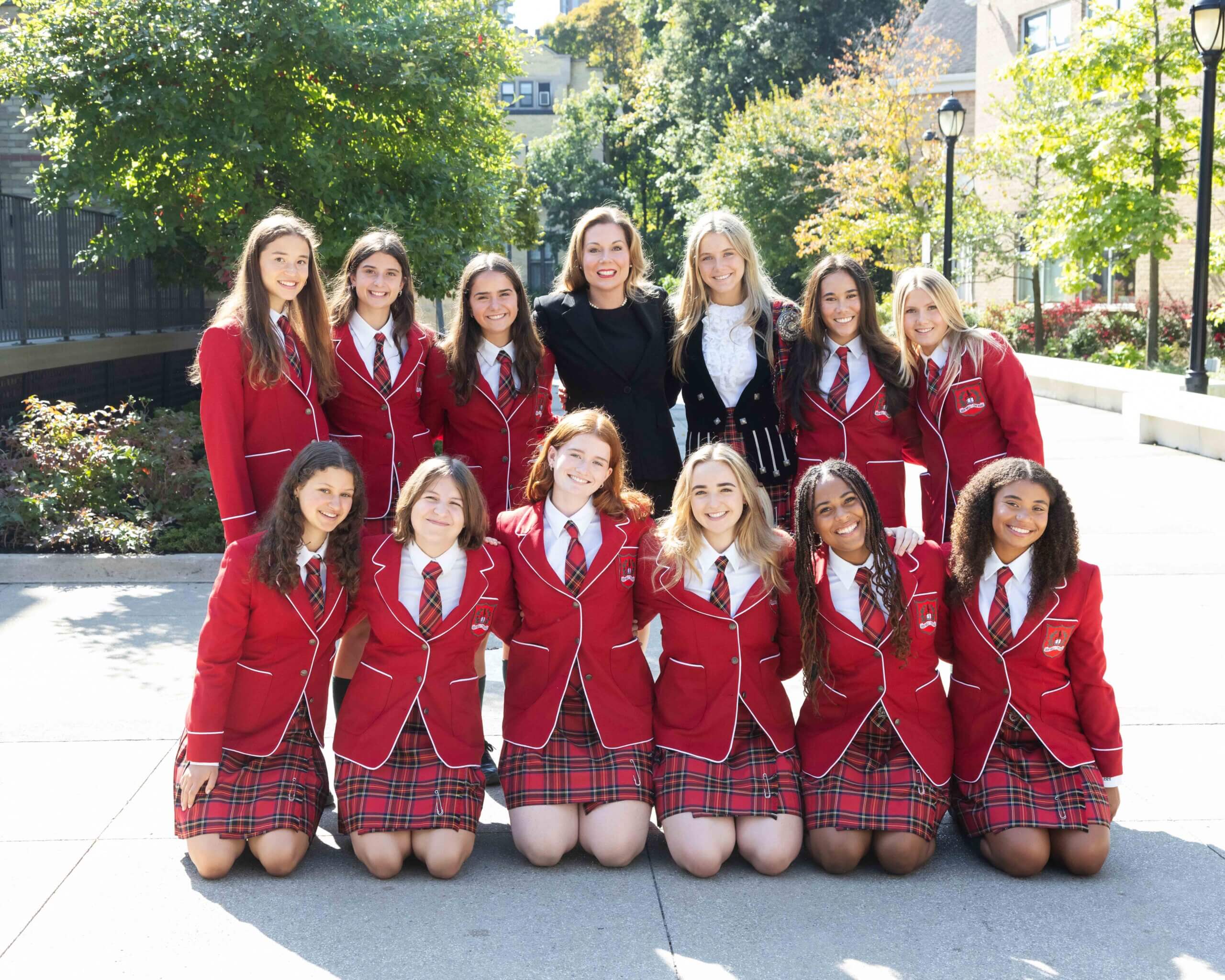 A group of thirteen smiling young women, most wearing matching red blazers and plaid skirts, pose together outdoors on a sunny day. Two adults stand in the back row among the group. Trees and buildings are visible behind them.