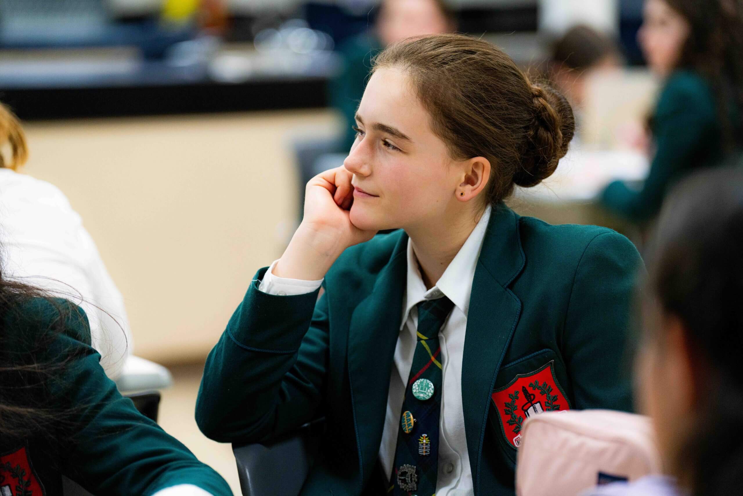 A student wearing a green school blazer sits at a desk, resting her chin on her hand and looking thoughtfully to the side in a classroom setting. Other students are blurred in the background.
