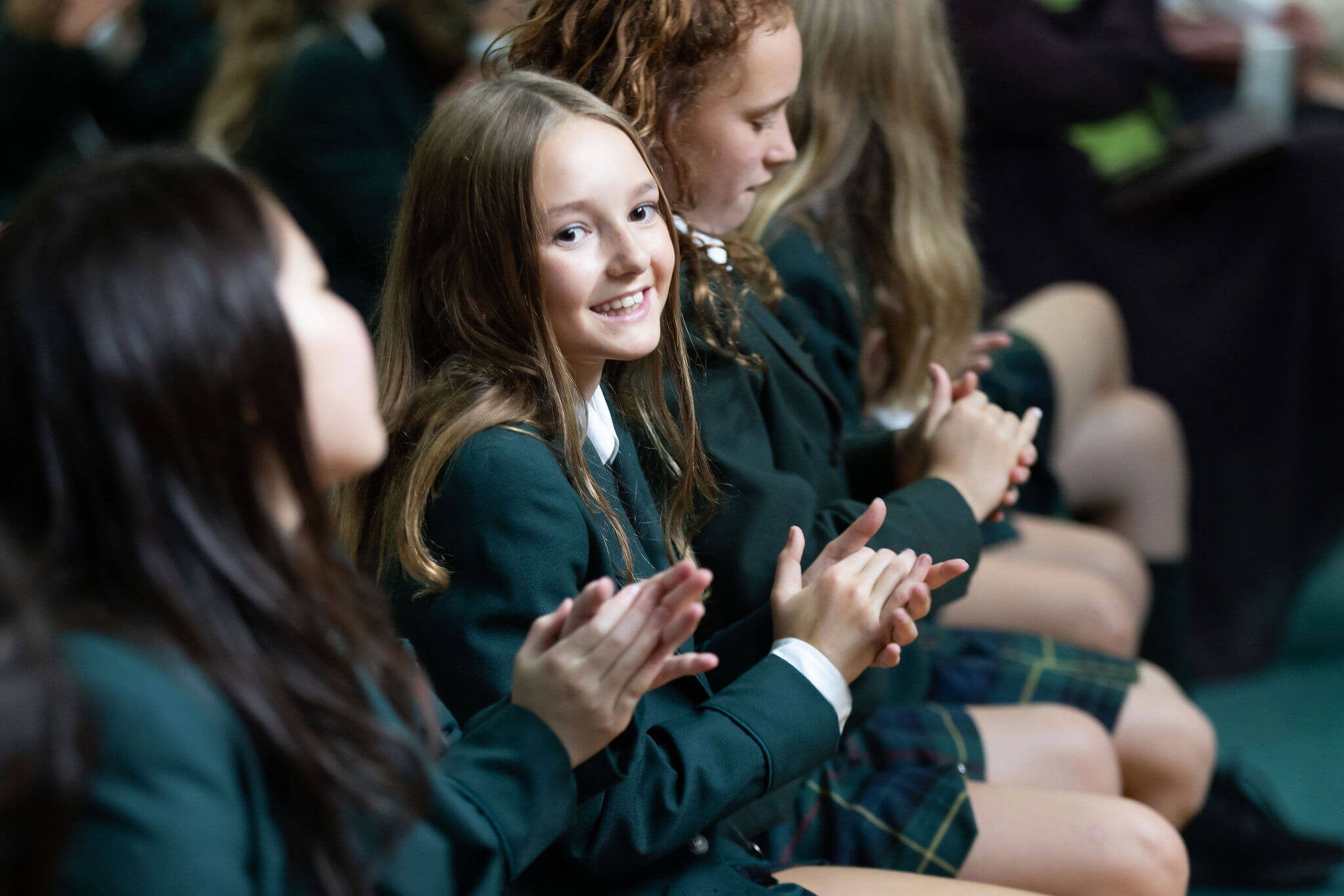 A smiling girl in a green school uniform claps while sitting in a row with other students, who are also clapping, at what appears to be a school event or assembly.