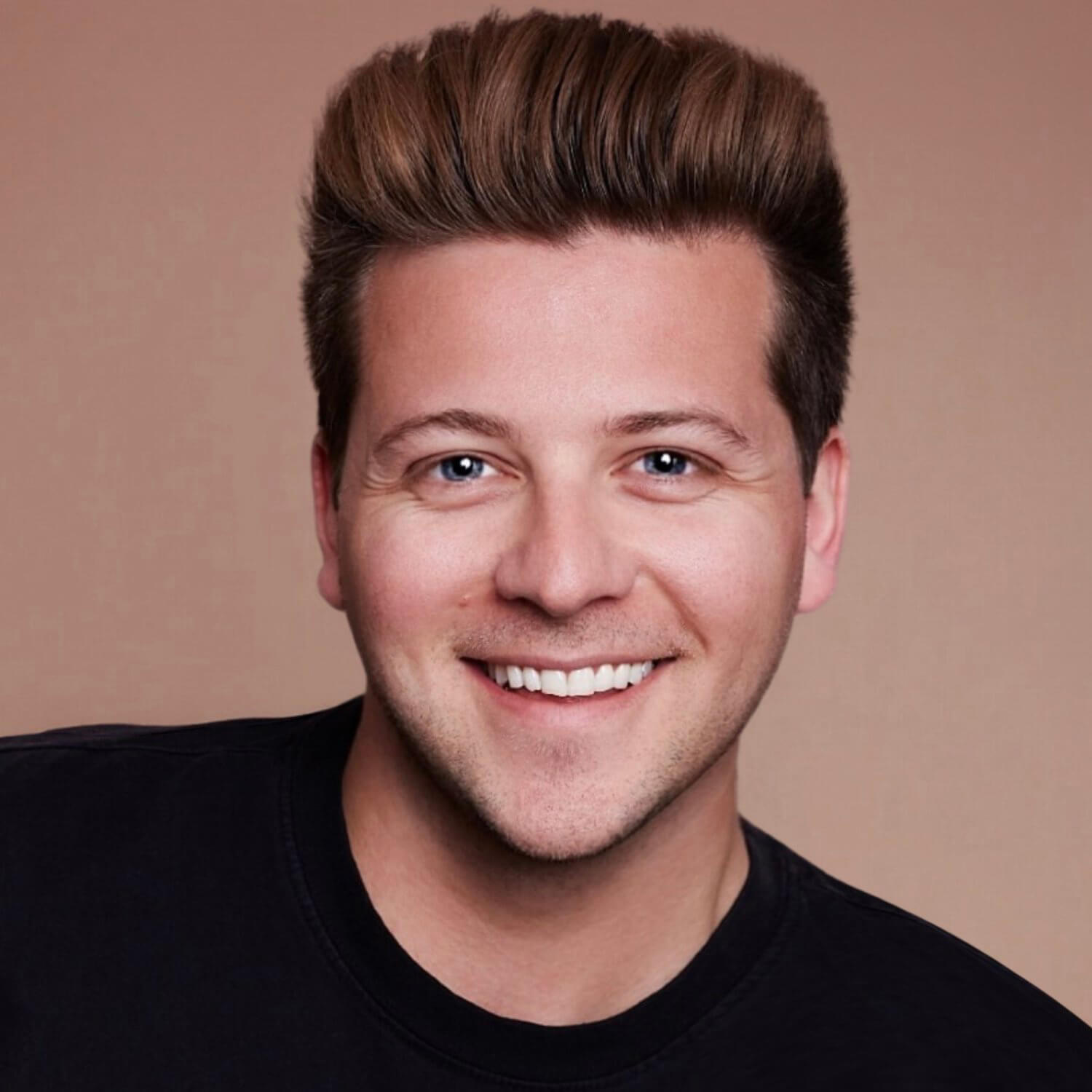 A smiling man with light skin, styled brown hair, and blue eyes wears a black shirt and poses in front of a plain, light brown background.