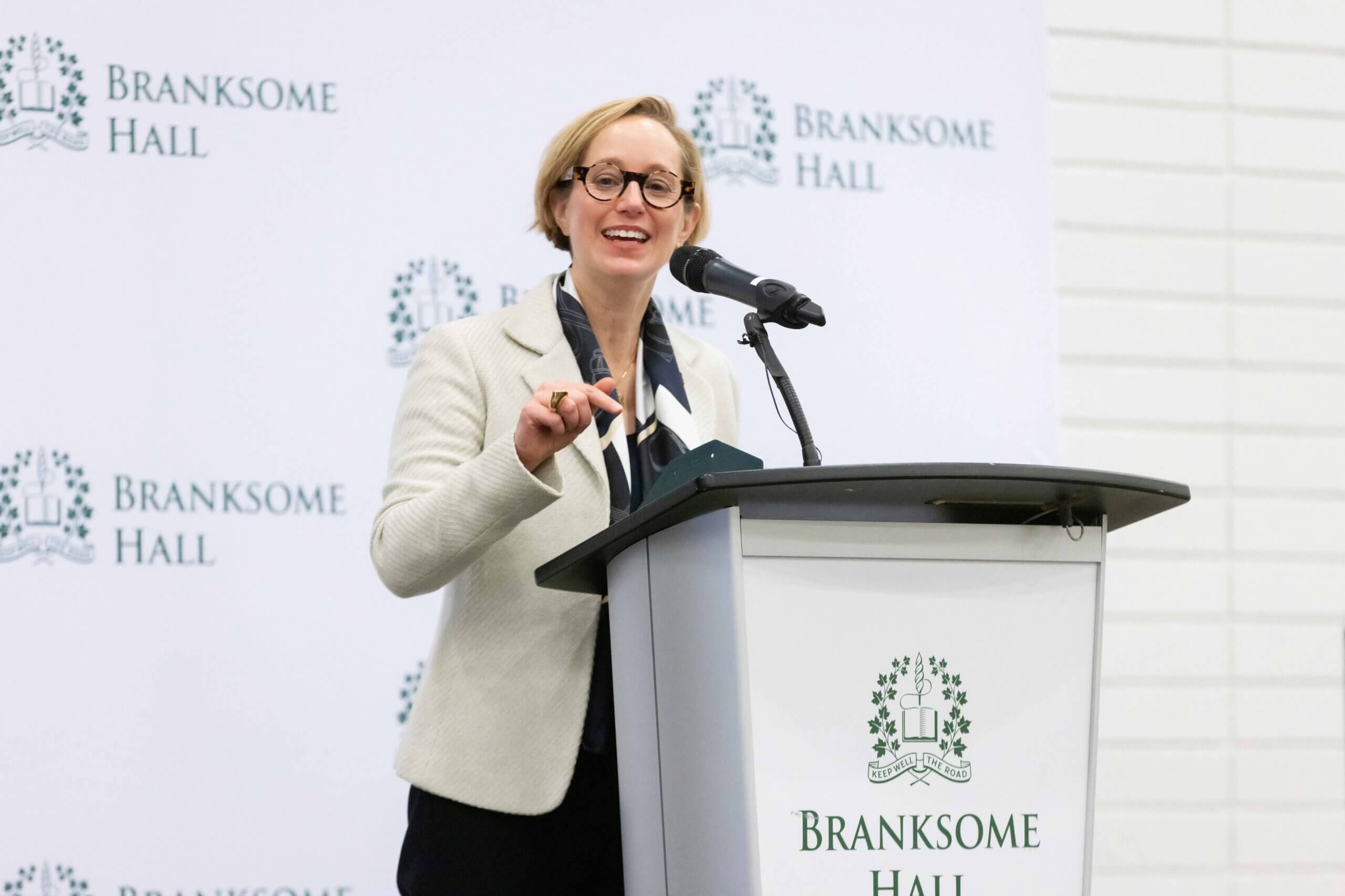 A smiling person wearing glasses and a light-colored blazer speaks at a podium with the Branksome Hall logo and backdrop, gesturing with one hand.