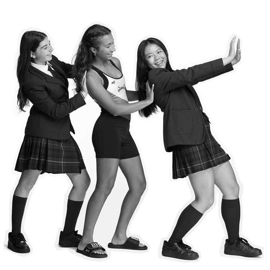 Three teenage girls pose playfully in a line. Two wear plaid skirts, blazers, and uniforms, while the middle girl wears a swimsuit and slides. They appear to be laughing and having fun. The background is white.