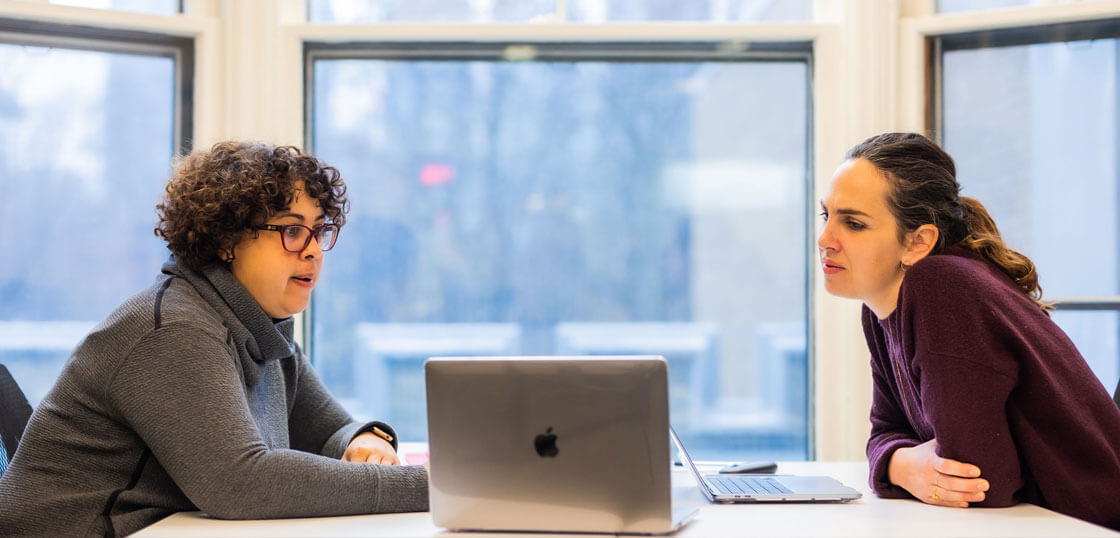 Two people sit at a table in front of a laptop, engaged in conversation. Large windows in the background show a blurry, wintry outdoor scene. One person wears glasses and a sweater, the other wears a dark top.